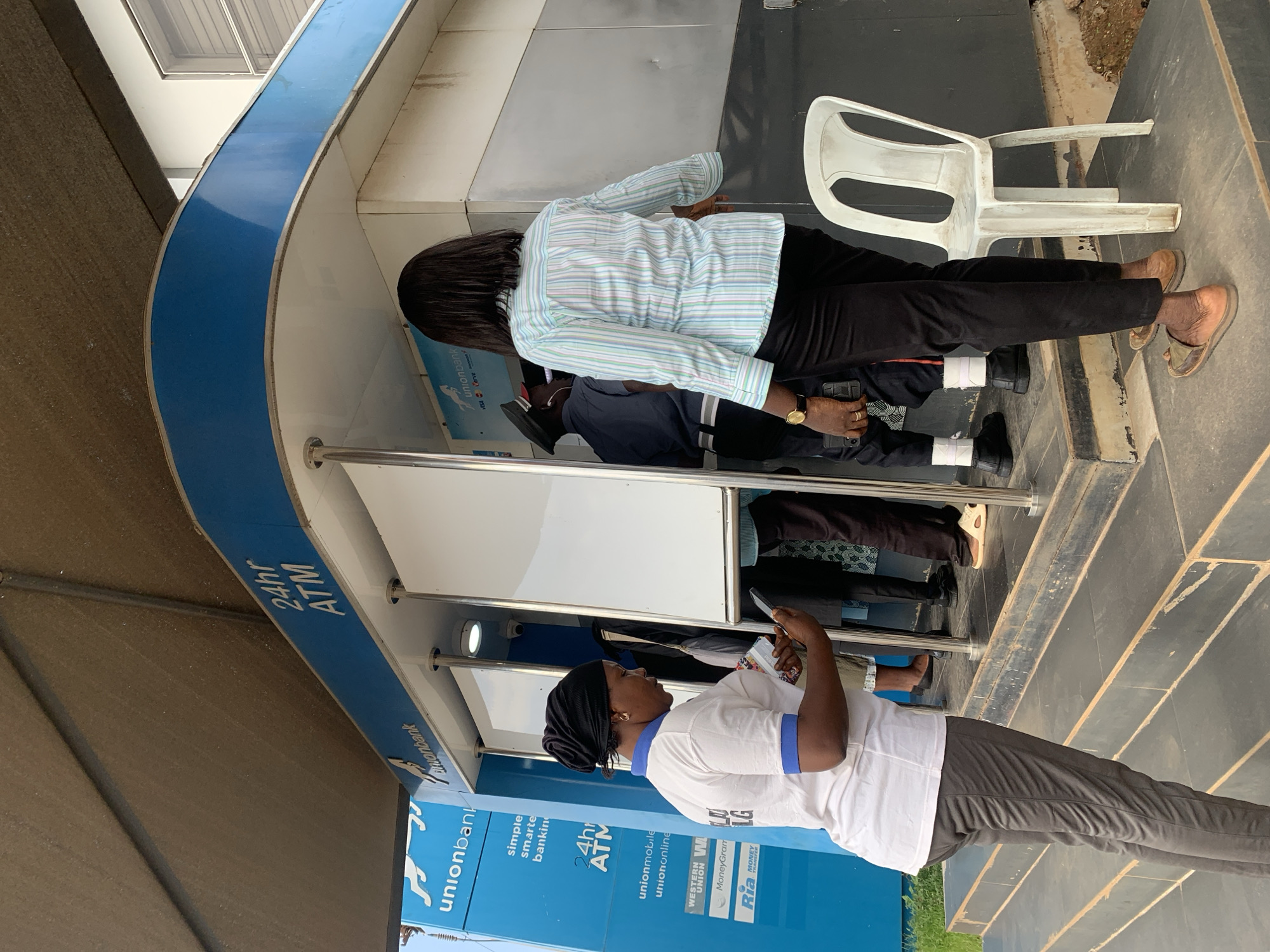 In the city of Jos, Nigeria, an ATM vestibule displays prominent "unionbank" signage and "24hr ATM" text. A woman wearing a white t-shirt with blue trim and a black headscarf stands to the left, looking at her smartphone. To her right, a woman in a striped shirt and black pants faces away from the camera, seemingly interacting with the ATM. Behind her, a security guard in uniform stands at an ATM. A white plastic chair sits empty to the right of the scene. The ground is paved with worn steps leading up to the vestibule.
