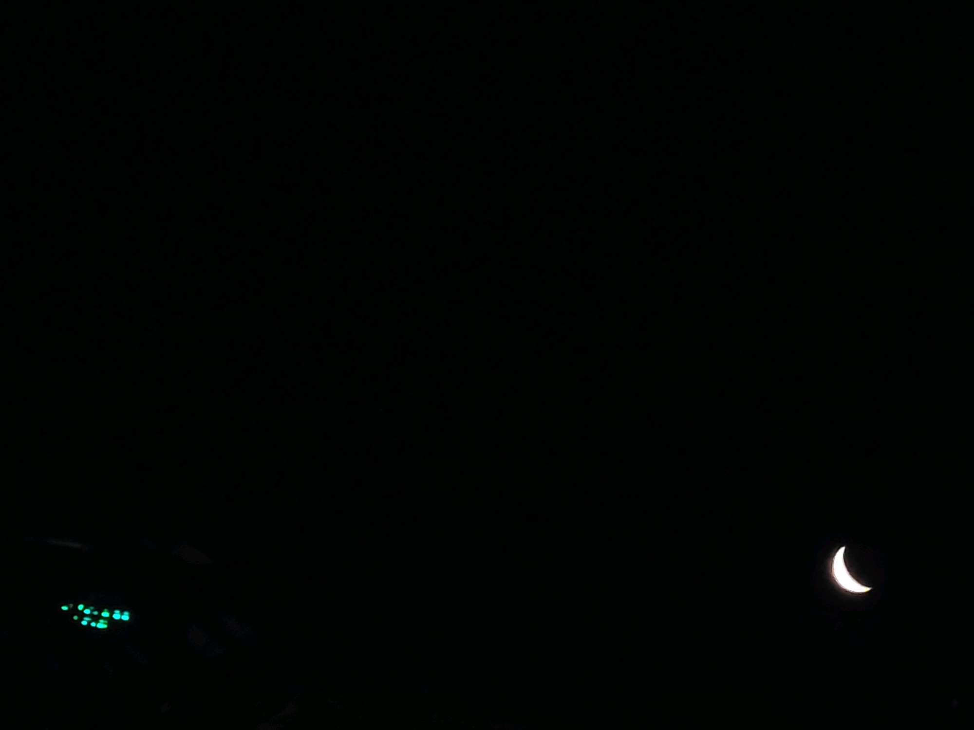 The image depicts a nighttime scene with a dark, starless sky. On the left side of the frame, a cluster of small, green lights is visible, possibly indicating a control panel or indicator lights on some equipment. To the right, a crescent moon shines brightly against the black expanse. The background is predominantly black, suggesting an outdoor setting away from significant light pollution. The overall impression is one of a quiet, dark night, with the moon being the most prominent celestial feature. There are no people or specific identifiable landmarks that would pinpoint the location within Ozolnieki, Latvia. The scene appears to be a static, observational shot.