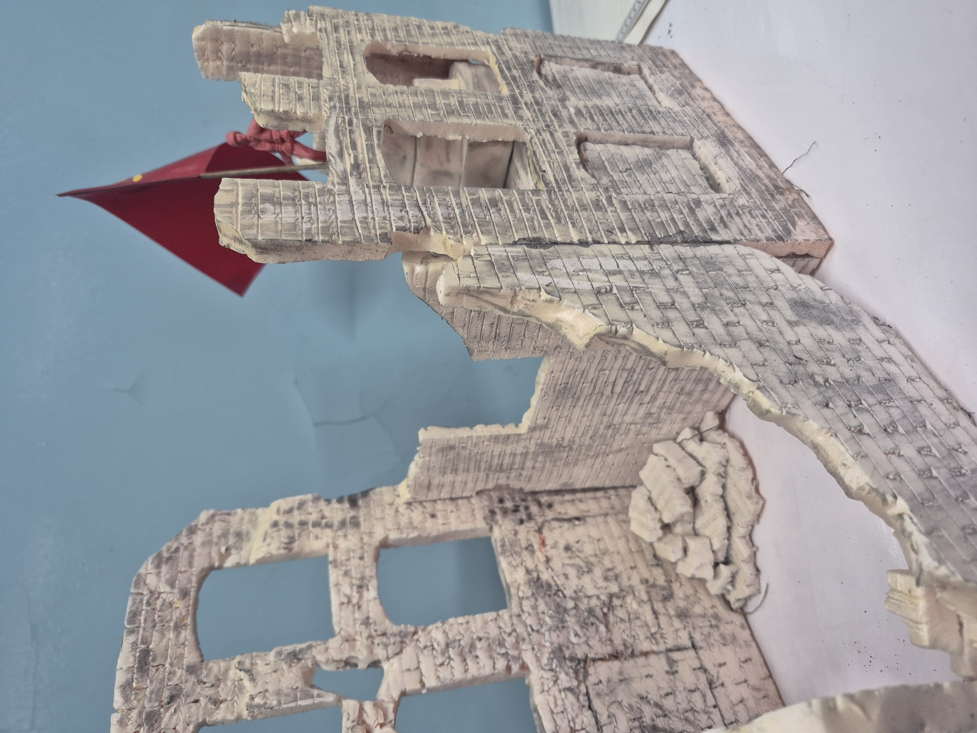 A diorama depicts the ruins of a brick building with arched windows against a light blue background. A small, red toy soldier stands on top of the ruins, holding a red flag with a yellow star. Debris made of lighter colored material is scattered within the ruined structure, suggesting destruction. The diorama is placed on a white surface, and the background appears to be a wall. There is no indication of the city of Chaikovsky, Russia, in the image.