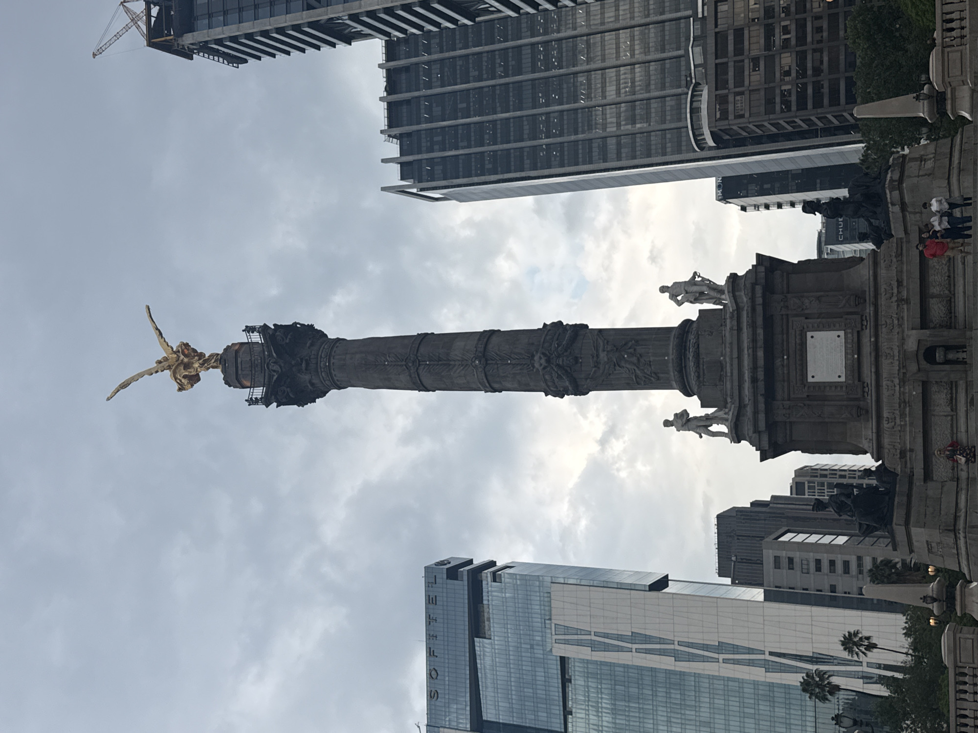 The image is a low-angle, vertical shot of the Angel of Independence monument in Mexico City, Mexico, framed by tall buildings and a cloudy sky. The monument's golden angel statue is atop a tall column. Statues and ornate carvings are on the base. Tall, modern buildings surround the monument, one with "SOFITEL" vertically displayed on its facade. A construction crane is partially visible above one of the buildings. Several people stand near the monument's base, observing the scene. The sky is overcast with a diffused light.
