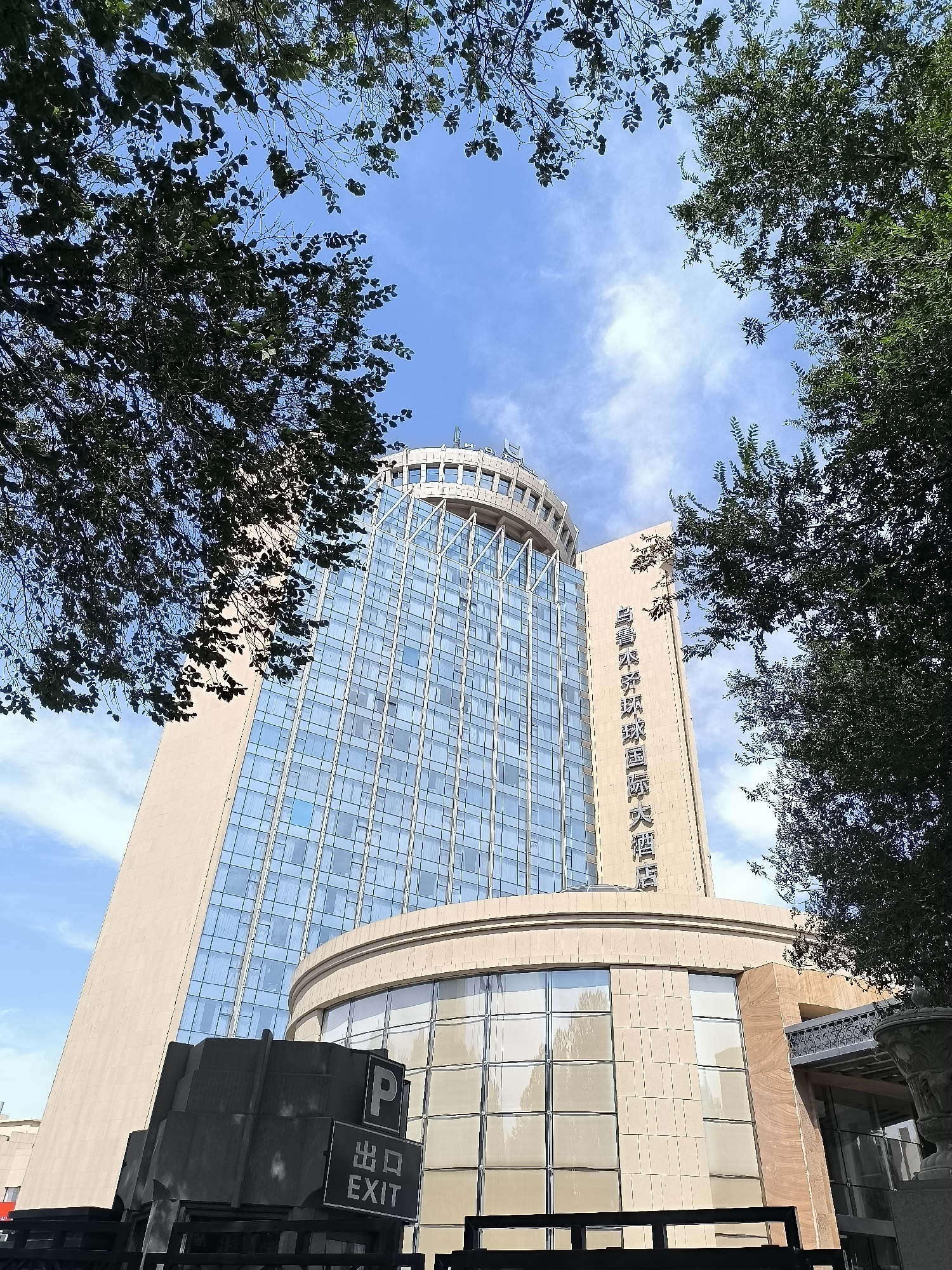 This is a low-angle shot of a modern hotel building in Urumqi, China, viewed from below through the branches of leafy trees. The building features a beige facade on the left and a glass facade on the right, with a curved section in the foreground also clad in beige material and large windows. Vertical Chinese characters are visible on the beige portion of the building, reading "乌鲁木齐君庭国际大酒店," which translates to "Urumqi Junting International Hotel." In front of the curved section of the hotel, a dark, industrial-looking structure is visible, with a "P" symbol indicating parking and a sign that reads "EXIT" in English and Chinese characters. The sky above is blue with scattered white clouds. The overall scene suggests an urban environment on a clear day.
