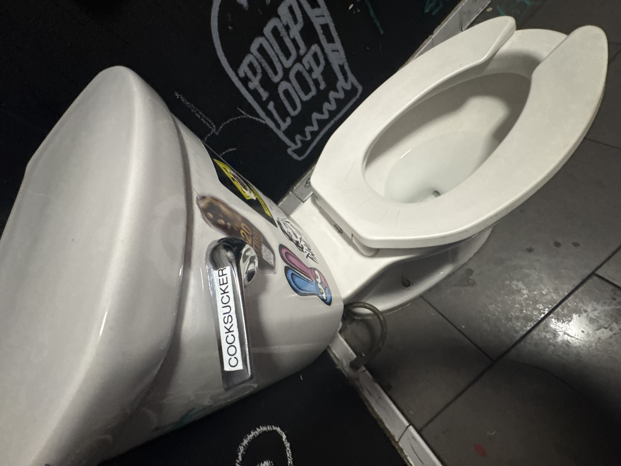 A toilet is photographed in a restroom. The toilet is white with an open seat and is adhered with stickers, including one that reads "COCKSUCKER". Graffiti with "POOP LOOP" is written on the wall. The floor tiles are gray and the surrounding walls are black. The location is Las Vegas, Nevada, USA.