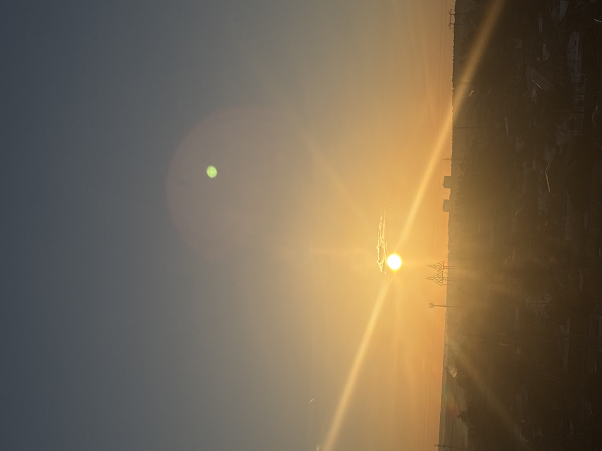 The image captures a sunrise or sunset scene in what appears to be Chaikovsky, Russia. The sky transitions from a dark blue to an orange-yellow hue near the horizon where the sun is situated. The sun itself is bright, with visible rays emanating from it. The skyline features the silhouetted forms of buildings, trees, and power line structures, suggesting an urban or industrial area. There's a soft, circular glare in the sky, possibly a lens flare.