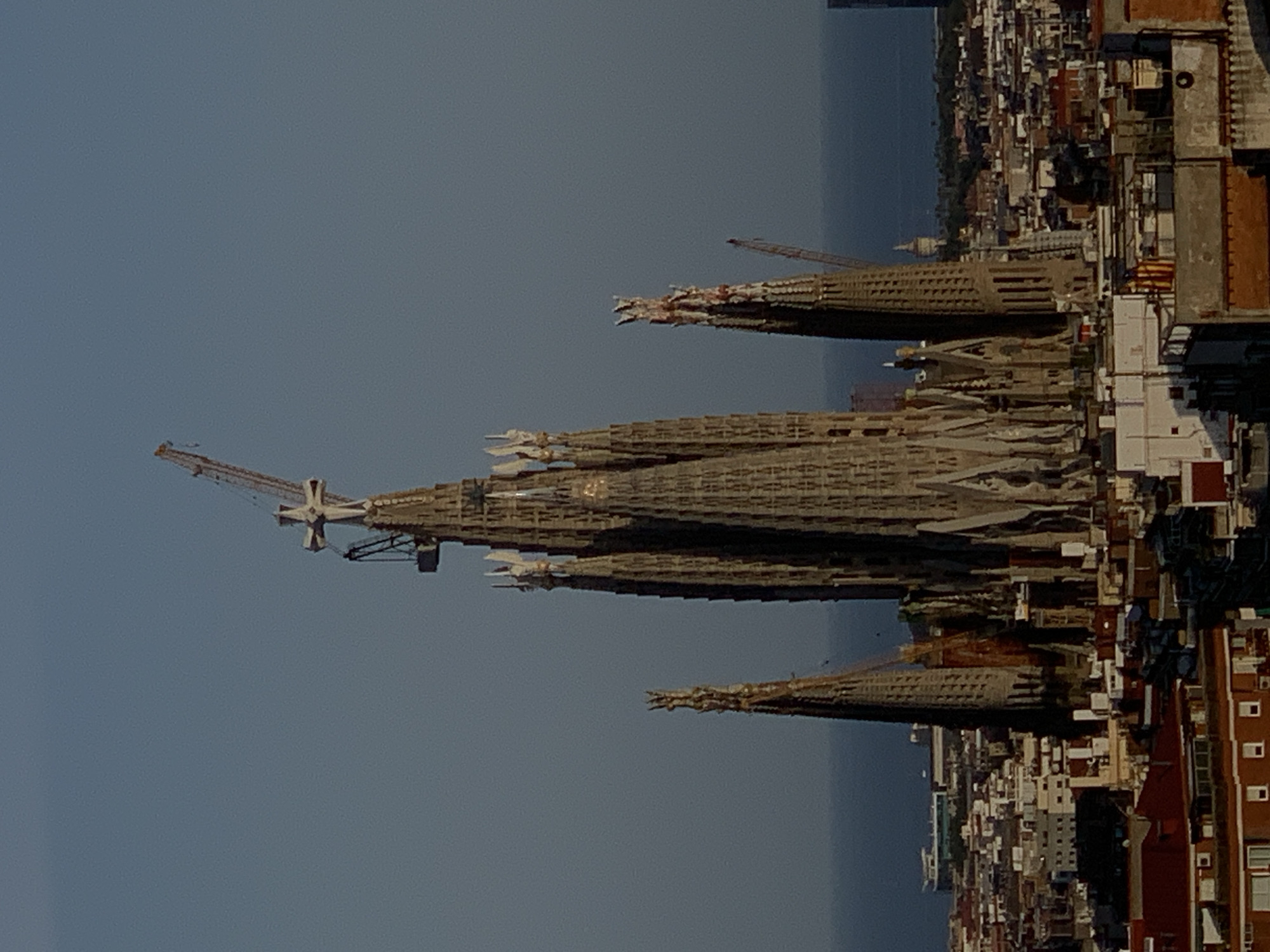 The image displays a panoramic view of the Sagrada Familia basilica in Barcelona, Spain. The basilica, a UNESCO World Heritage site, is a magnificent Roman Catholic minor basilica designed by Catalan architect Antoni Gaudí. The construction of the basilica is ongoing, as evidenced by the presence of construction cranes and scaffolding around the spires.
The basilica's distinctive architecture features intricate stone carvings, soaring spires, and stained-glass windows that cast colorful light within. The basilica is surrounded by a dense urban landscape of buildings, rooftops, and streets. In the background, the Mediterranean Sea stretches to the horizon, with a clear blue sky above. The overall impression is one of awe-inspiring grandeur and ongoing artistic creation.