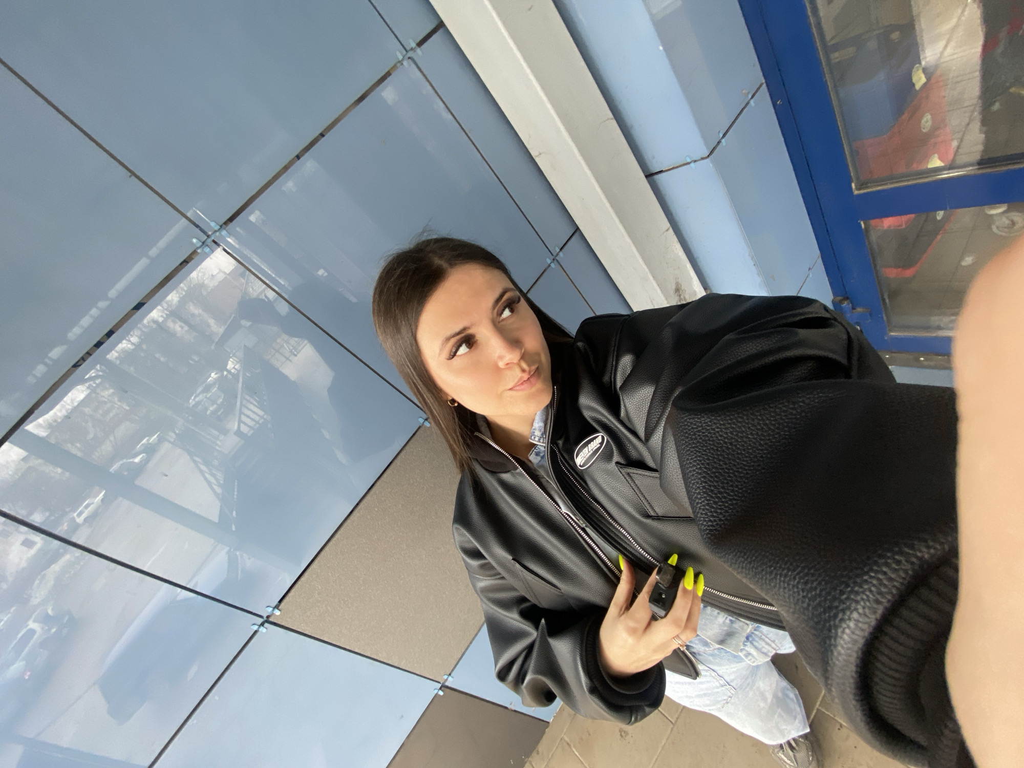 A young woman with dark hair and olive skin, wearing a black leather jacket and jeans, is standing under a tiled blue and grey ceiling, possibly in a public space in the city of Chaikovsky, Russia. The woman holds a black rectangular object in her hand, her nails are painted neon yellow. The photograph has a slightly rotated perspective. The surroundings include a wall with blue and grey tiled panels.