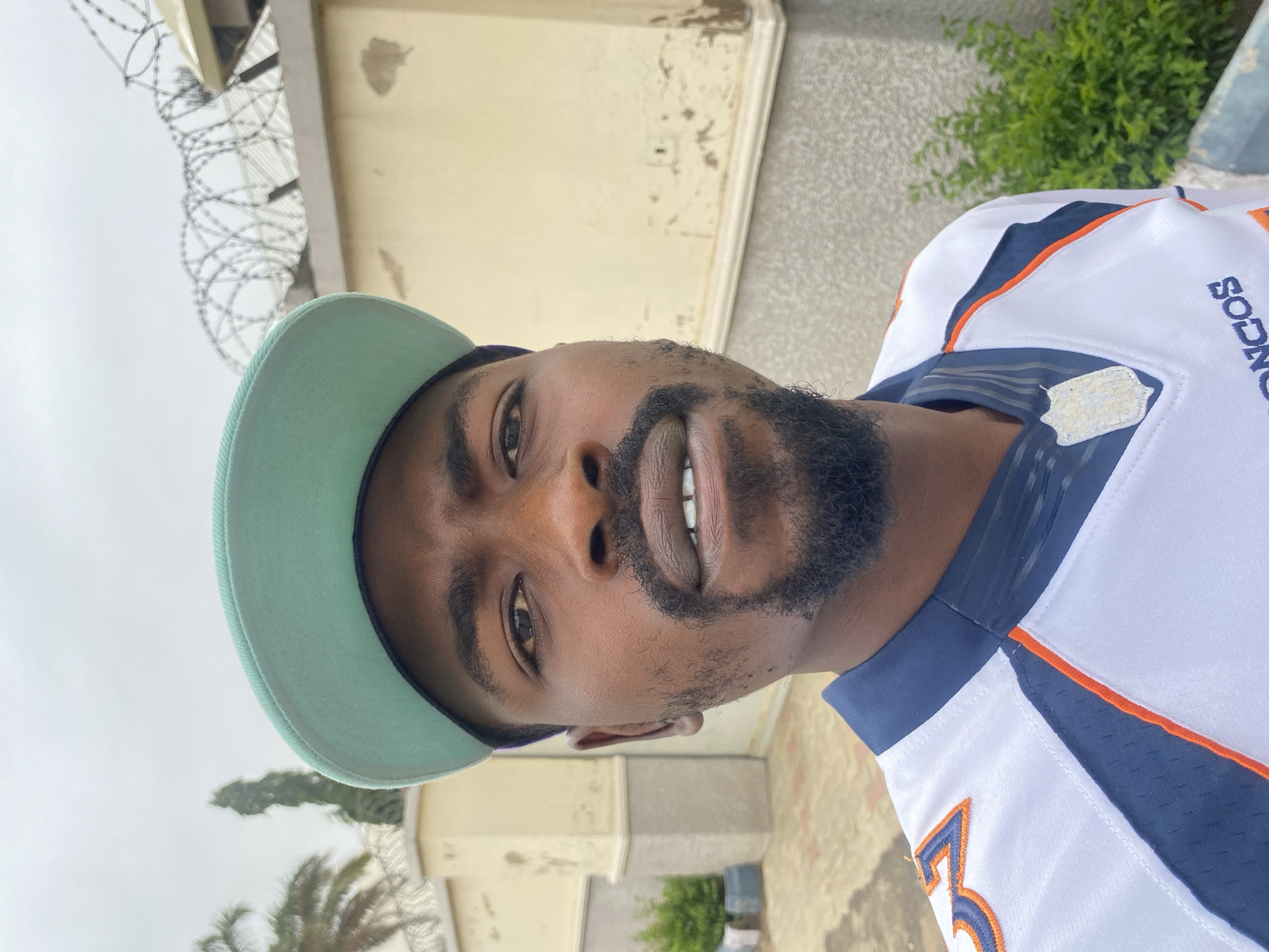 A close-up, vertically oriented portrait captured in Jos, Nigeria, features a Black man looking directly at the camera. He has a short, dark beard and is wearing a light blue or mint green baseball cap and a white sports jersey with navy blue and orange detailing.
The man's expression is calm and neutral, with a slight smile that reveals a portion of his upper teeth. The background is an overcast sky, indicating daytime under cloudy weather conditions. To his left, coiled barbed wire is visible atop a light-colored wall or fence, suggesting a secured area. Further back, buildings and green foliage, including what appears to be a palm tree, are partially visible, consistent with an outdoor, semi-urban setting.
Visible text on the man's jersey includes a partial vertical word "BRONCOS" and the number "3", suggesting it is a Denver Broncos American football jersey. The lighting is diffused and even, characteristic of an overcast day.