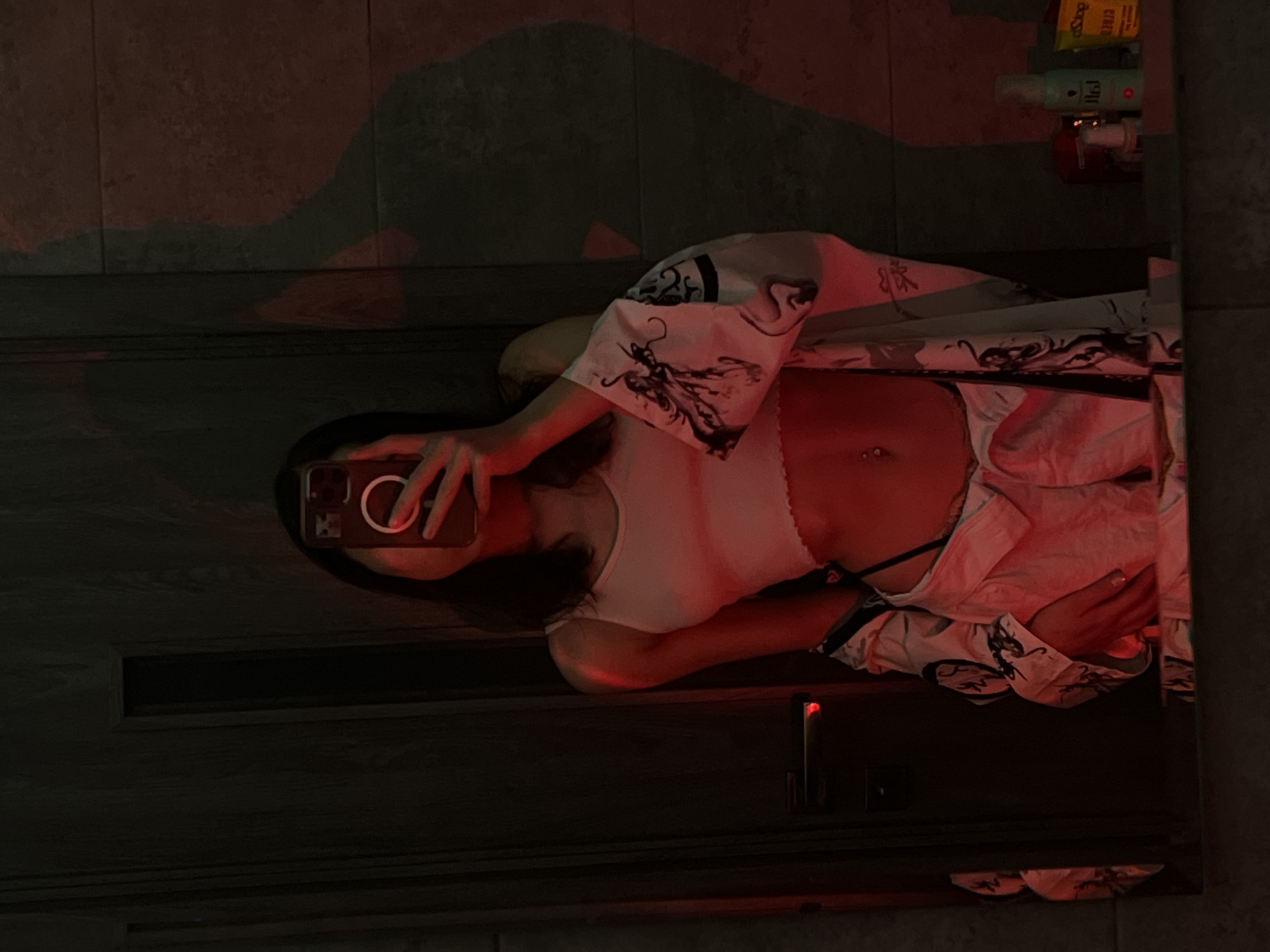 A person is taking a mirror selfie in a dimly lit room. The person is wearing a white tank top and a patterned robe with dragon designs. A phone is held up to the mirror to capture the reflection. The room appears to have tiled walls and a wooden door. There are some bottles of toiletries visible on a shelf in the background, reflected in the mirror. The lighting is red, casting a warm glow on the scene. The overall mood is intimate and relaxed. There is no visible text in the image.