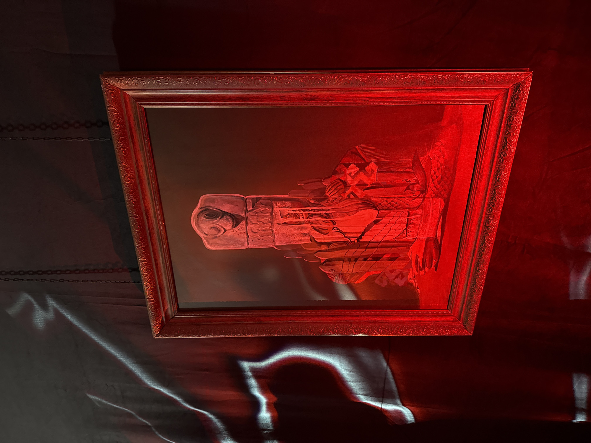 A framed, red-toned artwork hangs against a dark background, likely indoors. The artwork depicts a carved or engraved image featuring abstract and figurative elements. Light reflects across the artwork and the background. There is no clear indication of Yerevan, Armenia.