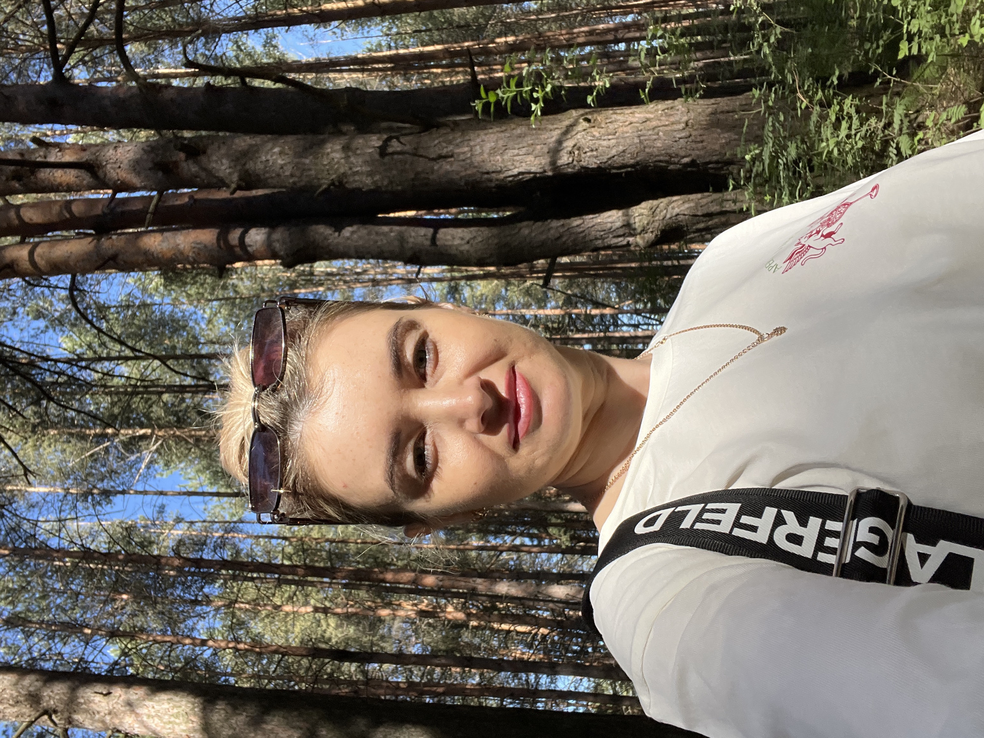 A woman is in a forest, possibly the city of Chaikovsky, Russia. She is wearing sunglasses on her head and a white shirt. The shirt has a "LAGERFELD" branded strap and a gold necklace. The background features tall trees and a blue sky visible through the branches. The image appears to be a selfie.