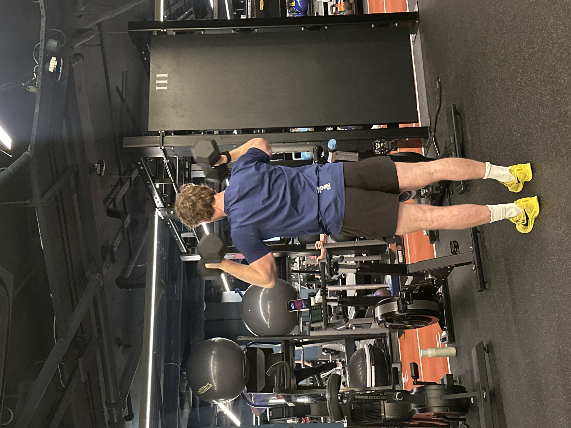 The image depicts a person in a gym setting. The person is wearing a dark blue shirt, black shorts, white socks, and yellow shoes, and is lifting dumbbells with both hands. The gym has various equipment including exercise balls, rowing machines, and a weight rack labeled "III". The floor is a combination of black rubber and a section with red lines, suggesting an area for running or sprints. The location in the city of Unknown, Unknown is unknown.