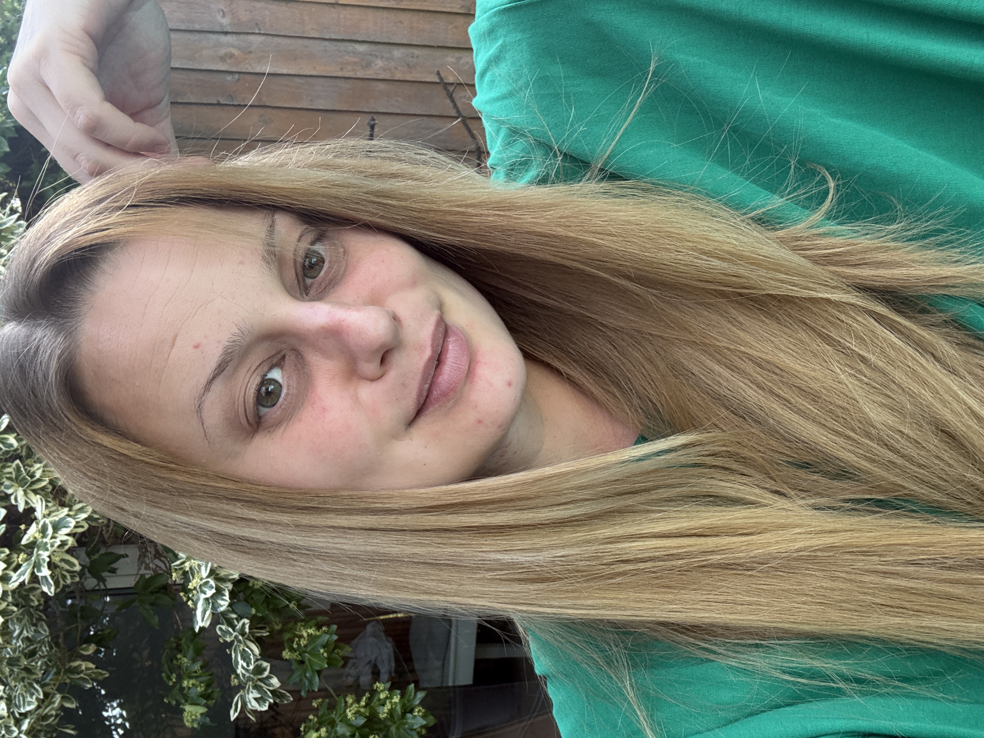 A blonde-haired woman with fair skin is photographed outdoors. She wears a green top, and her hair flows to the side, partially obscuring the background. She is looking directly at the camera with a slight smile. The background contains foliage and a wooden fence. The location appears to be Hazlemere, United Kingdom, based on visual details.
