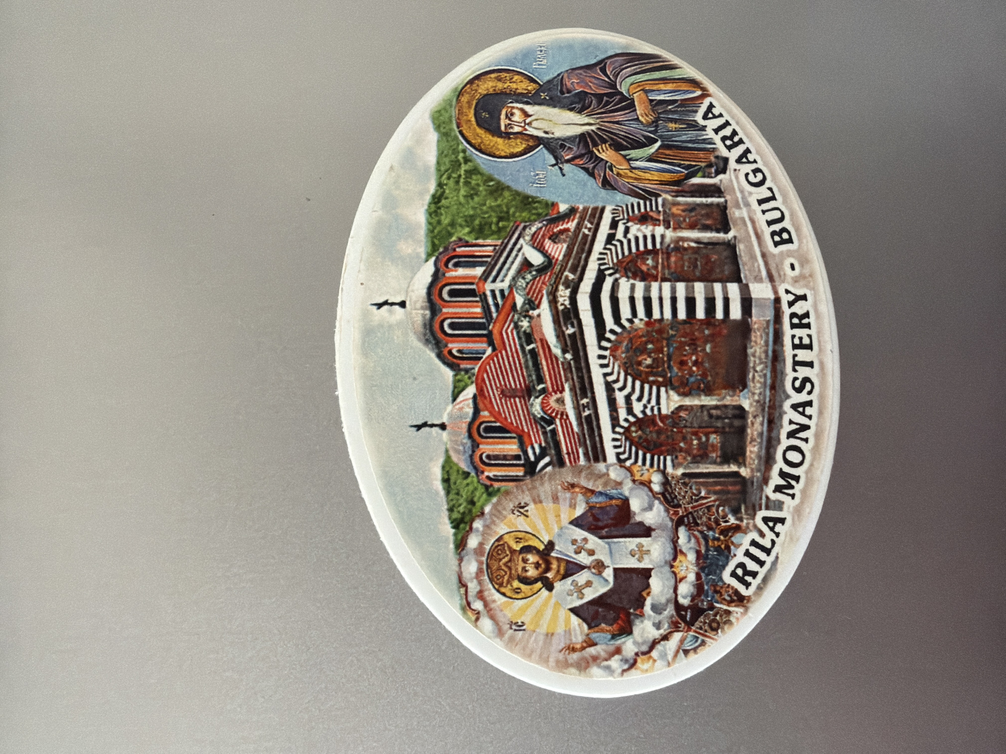 The image is a close-up of an oval-shaped magnet or sticker with an illustration of the Rila Monastery in Bulgaria. The illustration features a central depiction of the monastery's distinctive architecture, characterized by its striped arches and domed roofs, set against a backdrop of green hills. On the left side of the illustration is an image of a religious figure, possibly Christ, depicted with a halo and ornate robes. On the right side, another religious figure, likely a monk or saint, with a long white beard and a dark habit, is shown. Text at the bottom of the illustration reads "RILA MONASTERY - BULGARIA". The magnet is placed on a plain, reflective gray surface, likely a refrigerator door. The image does not depict the city of Pazardzhik, Bulgaria, as the Rila Monastery is a separate historical site located in the Rila Mountains.