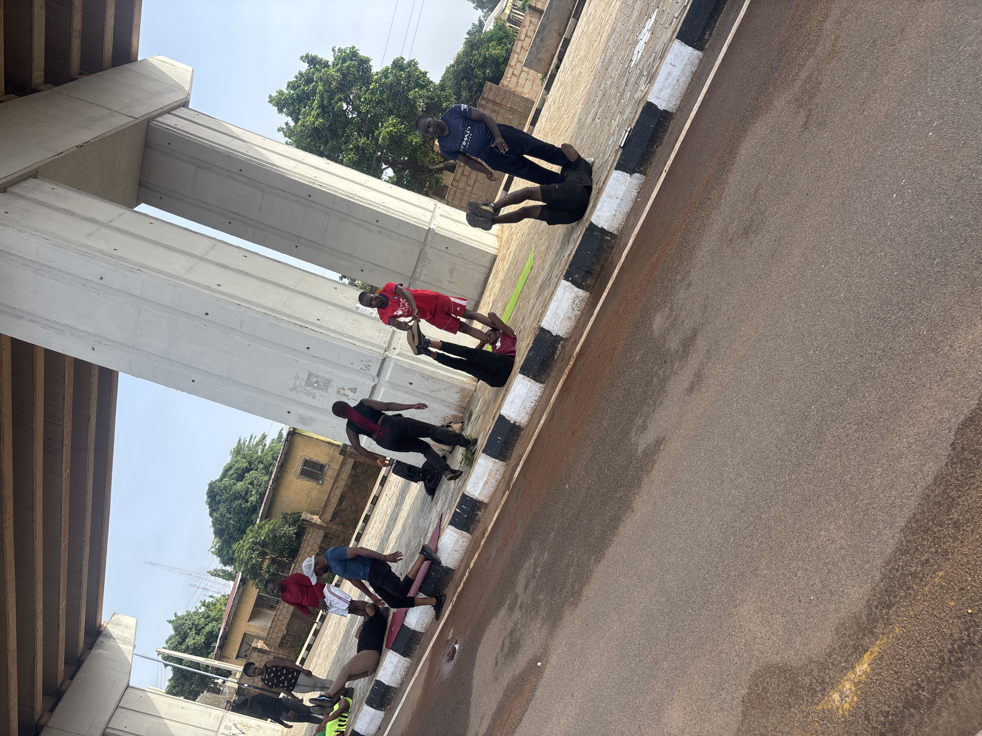 The image depicts an outdoor fitness session taking place under a large concrete overpass, likely part of a road or bridge infrastructure, in Jos, Nigeria. Several individuals are engaged in various exercises on the paved ground beside a road. One person is lying on their back with legs raised, while another is in a similar position, possibly performing sit-ups or core exercises. Two individuals are standing, one in a red shirt and shorts, and another in a blue t-shirt and black pants, observing or participating. Another person in black leggings and a maroon top is also on a green exercise mat. In the background, there are buildings with brown brick walls and yellow painted facades, and lush green trees. The sky is clear and bright. The overall scene suggests a community fitness group or personal training session.