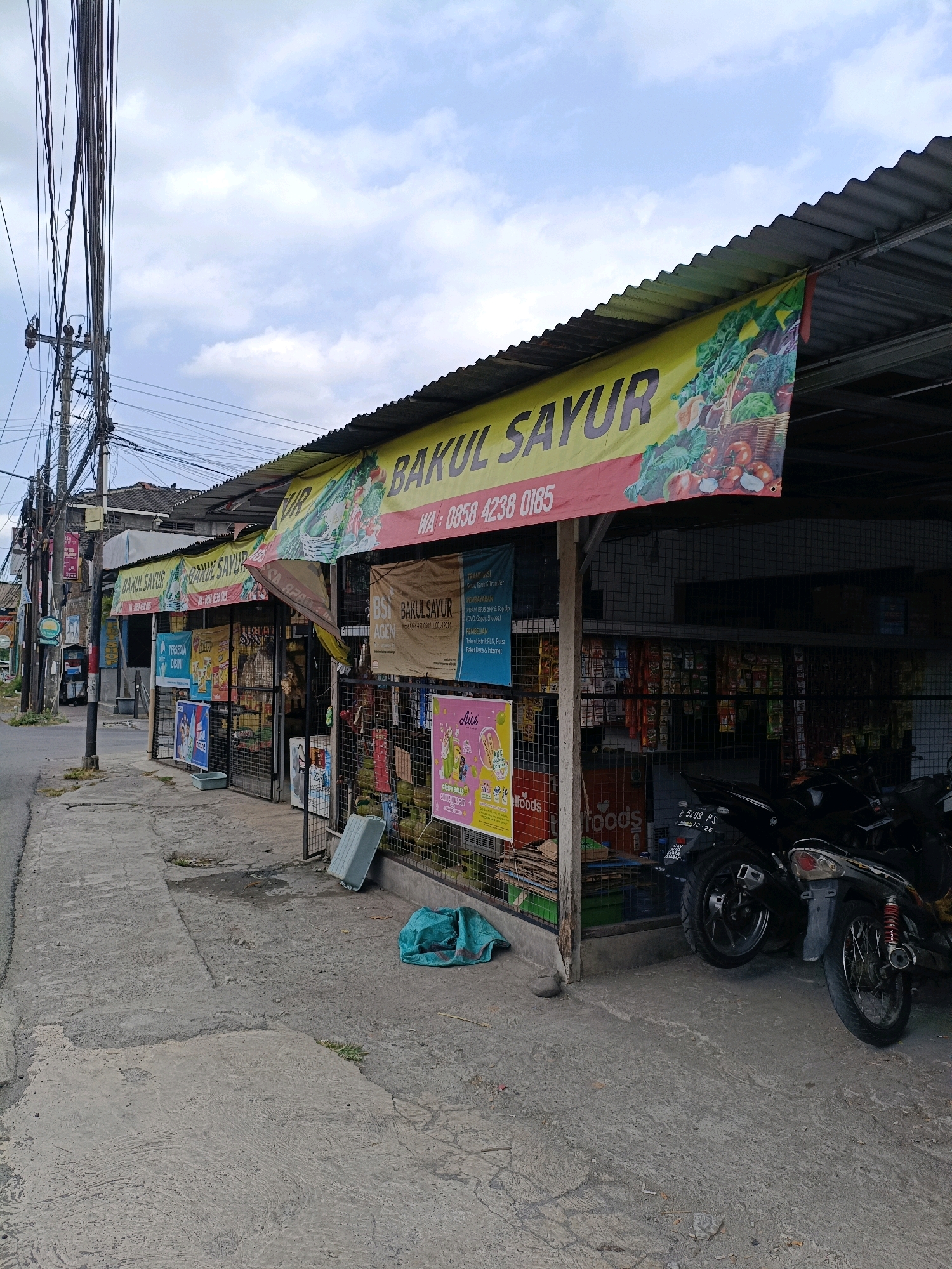 The image shows a street scene in Maguwoharjo, Indonesia, featuring a roadside vegetable stall. The stall is covered by a corrugated metal roof with a yellow banner displaying "BAKUL SAYUR" and an image of vegetables. Various advertisements and signs are hung on the stall's facade. In front of the stall, a sidewalk made of concrete runs alongside the street. To the right, two motorcycles are parked. Several green bags and a light blue plastic container are visible on the sidewalk near the stall. Electrical poles and overhead wires are prominent on the left side of the image, indicating an urban or semi-urban environment. The sky is partly cloudy.