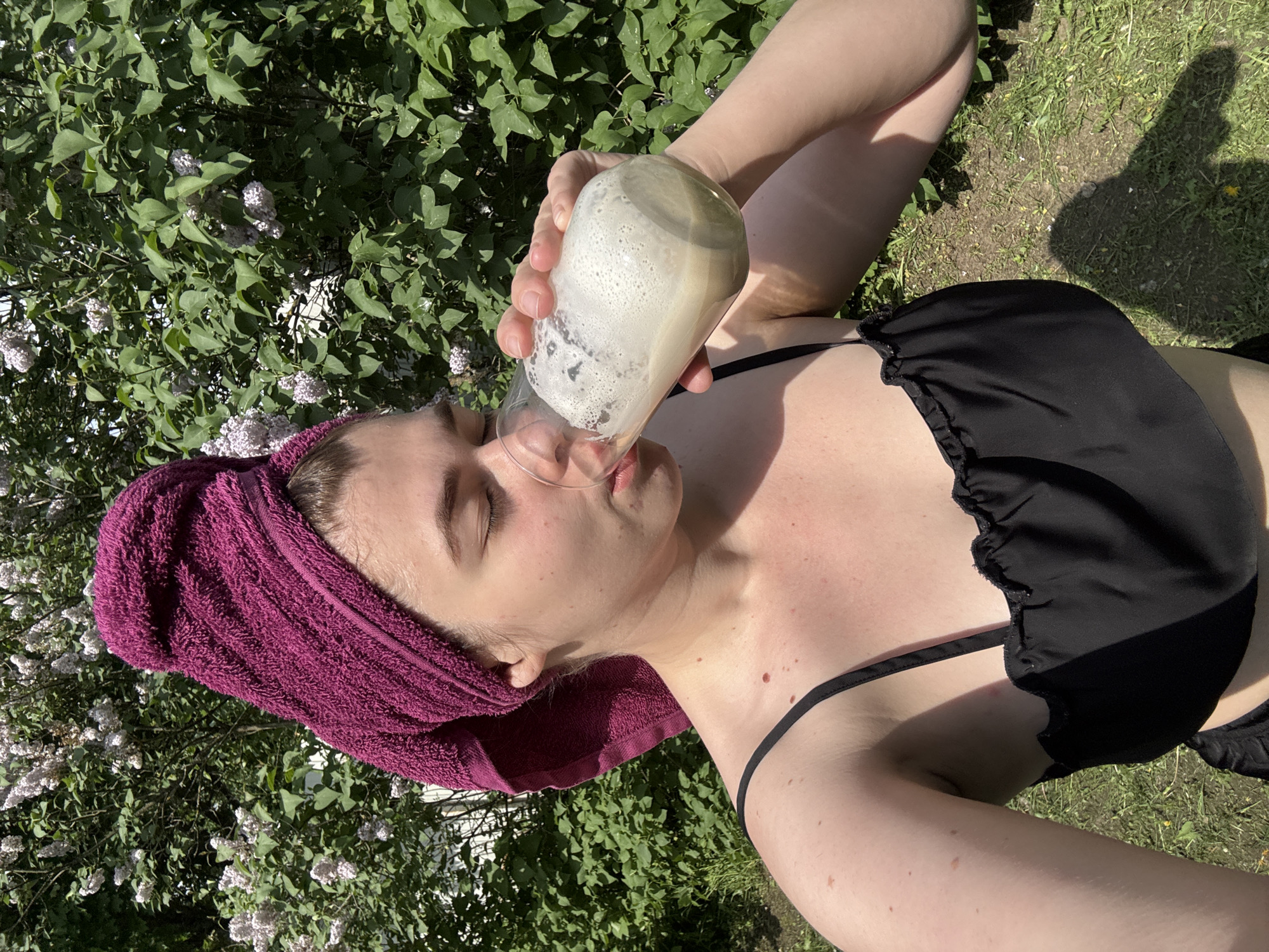 A woman lies on the grass wearing a black bikini and a purple towel wrapped around her head. She is drinking from a glass filled with a foamy beverage. She is located near a lilac bush. The location is not identifiable as Мещёры, Russia from this image.