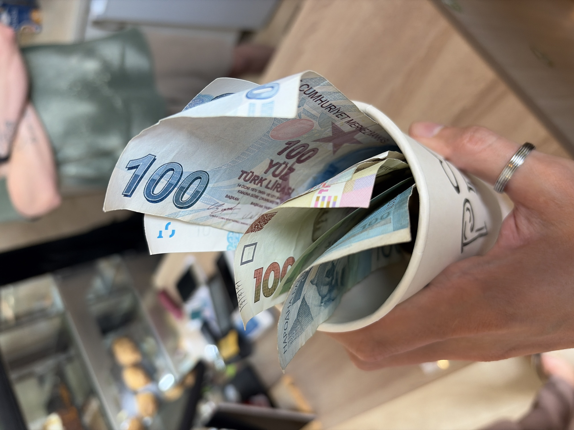 The image shows a person holding a white cup filled with a mix of Turkish Lira and possibly some Montenegrin banknotes. The Turkish Lira bills visible are 100 Lira notes. The person is wearing a silver ring on their finger. The background suggests an interior setting, perhaps a cafe or shop, with a display case containing food items visible in the blurred foreground. The style of banknotes suggests this may be in a tourist area. However, there is no information in the picture to confirm it is the city of Budva, Montenegro.