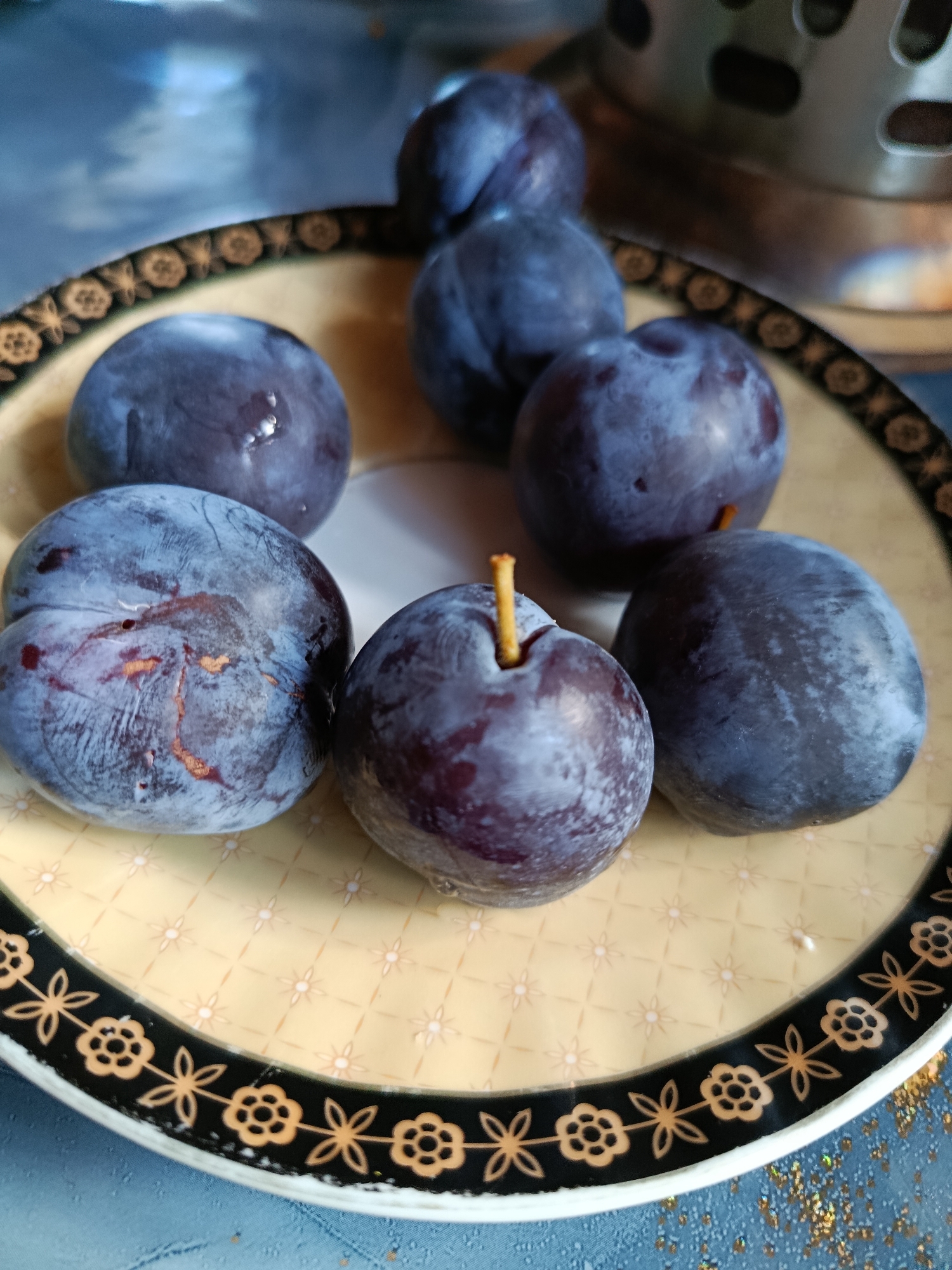 A close-up, top-down view shows seven plums on a light yellow, patterned plate with a decorative black and gold rim. The plums are deep purple with some lighter blue or gray dusty bloom. One plum in the foreground has a visible stem. The plate rests on a textured, blue surface, with a glimpse of a metallic object with holes in the blurred background. The image is well-lit, highlighting the textures of the fruit and plate. The location is Unknown, Unknown.