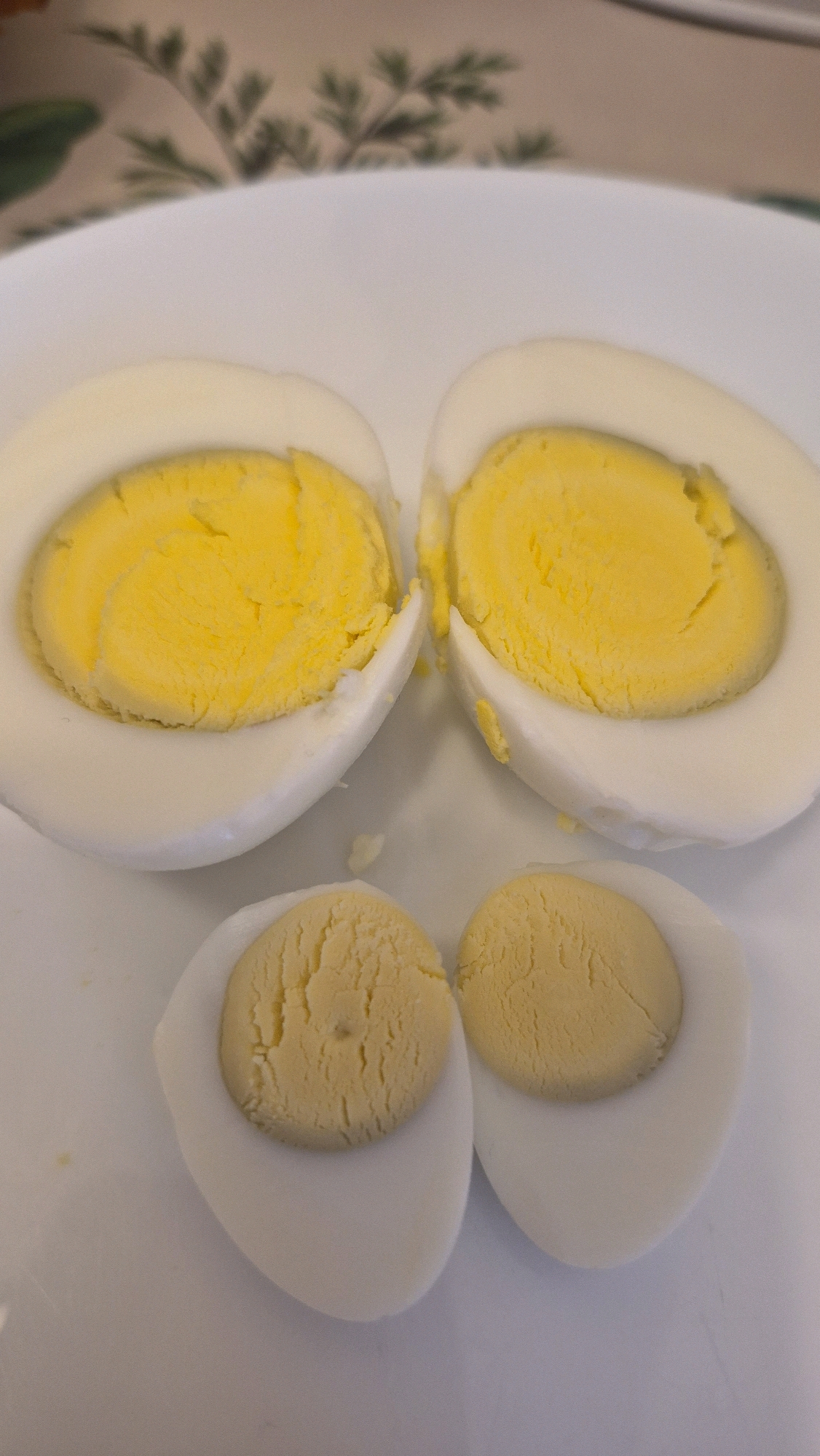 The image shows four halves of hard-boiled eggs arranged on a white plate. Two larger halves, containing bright yellow yolks and white egg whites, are positioned at the top of the frame, slightly apart. Below these, two smaller halves, each with a paler yellow yolk and white egg white, are placed side-by-side. The background is blurred, showing hints of a patterned tablecloth with green and brown elements. There are no people visible in the image. The scene is likely indoors, possibly in a kitchen or dining area. The image does not contain information about the city of Krauja, Latvia.