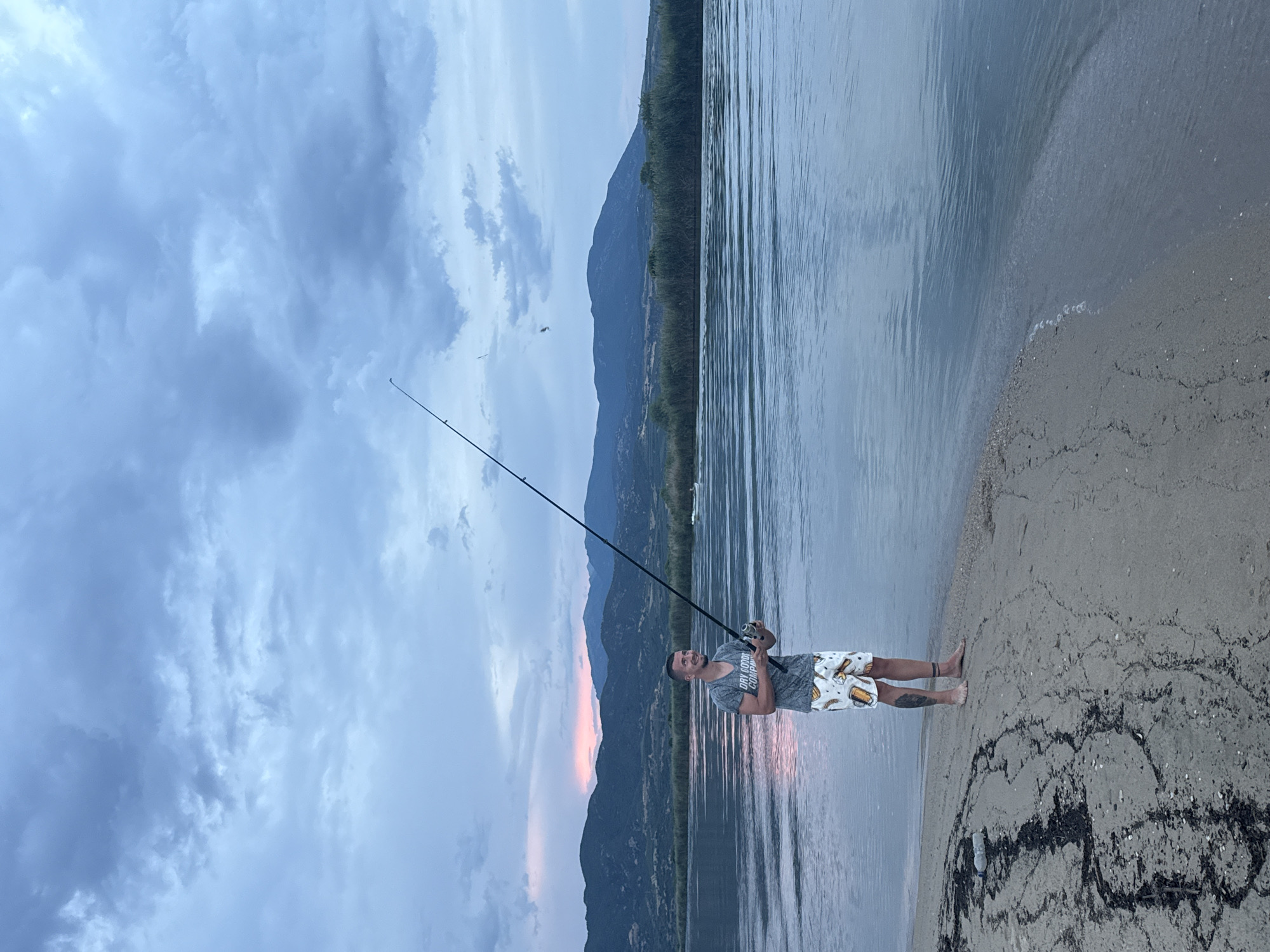 A man is fishing on the shore of a body of water, with mountains in the background. The sky is cloudy and there are hints of pink and orange from a sunset on the horizon. The man is wearing a grey t-shirt and patterned shorts, and he is holding a fishing rod. The water is calm, and there is a sandy beach in the foreground. There is also some debris on the beach, including a plastic bottle and seaweed. The overall scene is peaceful and serene.