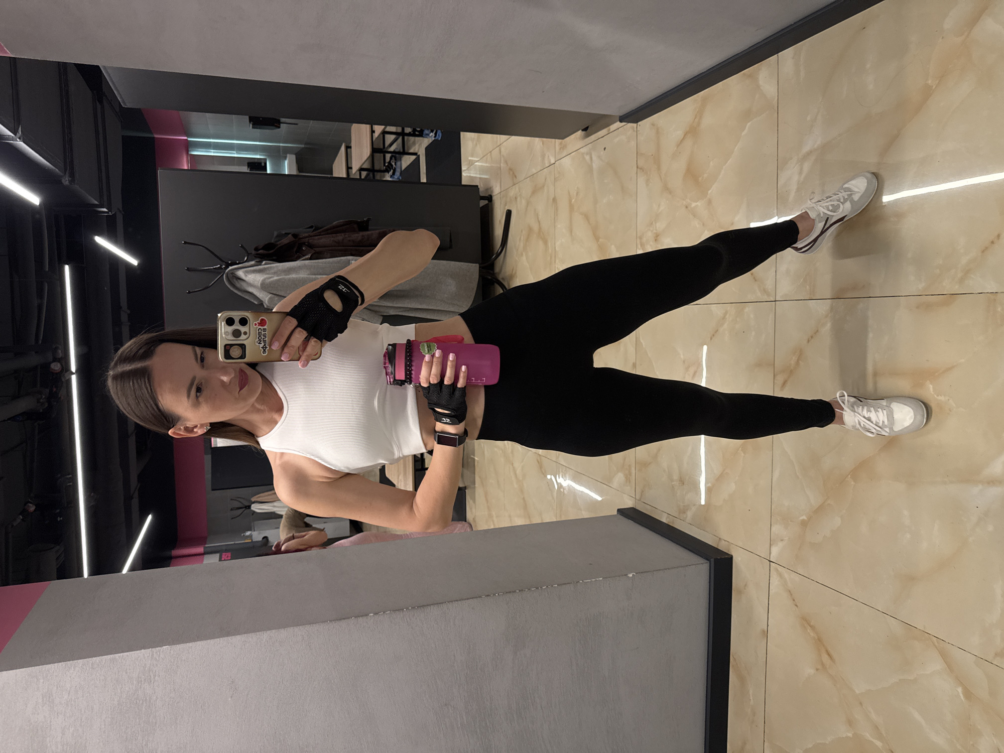 A woman takes a selfie in a gym setting. She is wearing a white tank top, black leggings, and black weightlifting gloves. She holds a pink water bottle. The gym has a modern design with black and pink accents and light marble floors. The image may have been taken at a gym in Sochi, Russia.