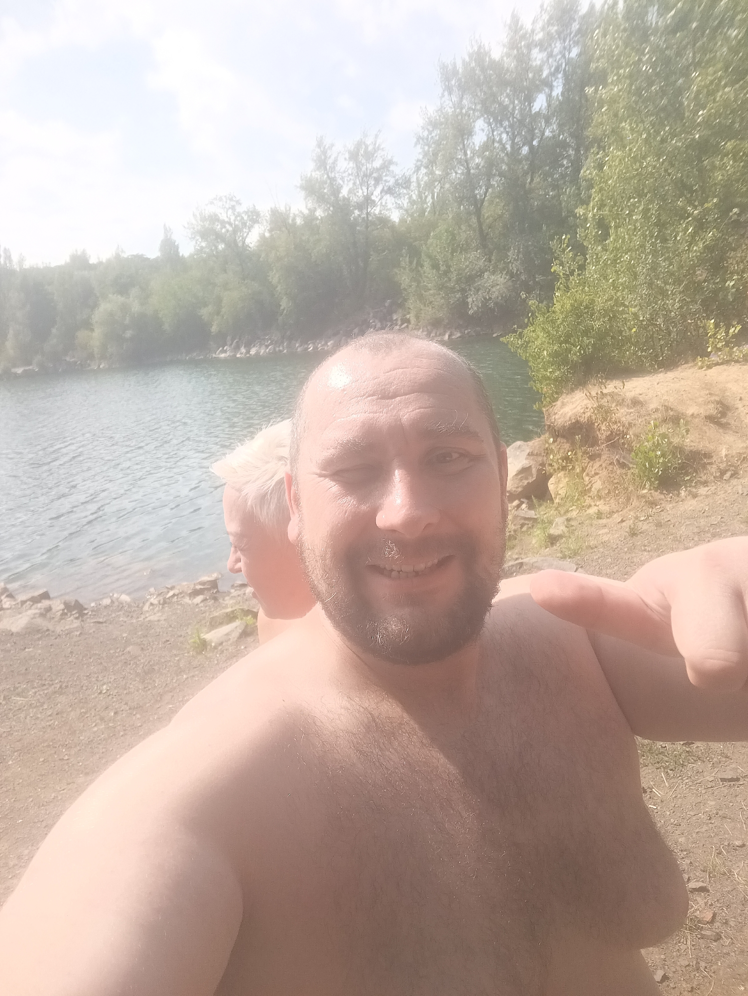 Two individuals are captured in a selfie taken outdoors by a body of water, likely a quarry given the rocky surroundings and clear water. The person in the foreground, a male with a beard and receding hairline, is smiling and looking at the camera. He is shirtless, as is the second individual, whose face is partially visible behind him. This second individual appears to have lighter hair. The background shows a shoreline with sand, rocks, and trees, suggesting a natural or semi-natural environment. The sky is bright and overcast. There is no indication of this specific location being within the city of Uzhhorod, Ukraine, or any other city.
