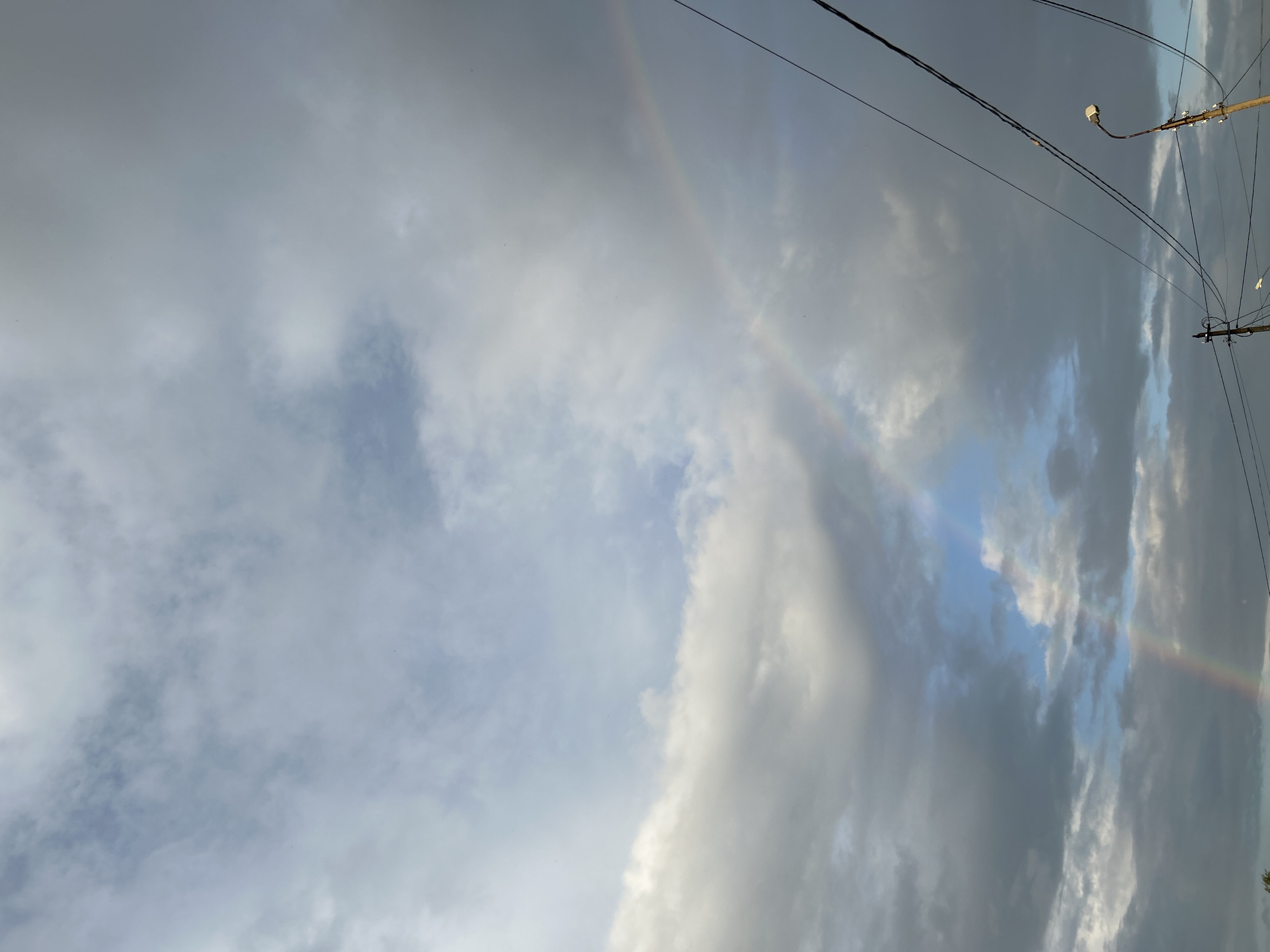 The image captures a sky scene in Chaikovsky, Russia, featuring a dramatic sky with a double rainbow arching across it. The clouds are varied, with some dark and heavy, suggesting recent or impending rain, interspersed with patches of lighter, brighter sky. The rainbow is most prominent in the middle right of the frame, with a fainter secondary rainbow visible below it. In the lower right quadrant, a street lamp on a pole is visible, with several dark power lines crisscrossing the sky. The overall impression is one of a post-rain atmosphere, with the natural phenomenon of a rainbow creating a colorful contrast against the moody clouds and man-made infrastructure.