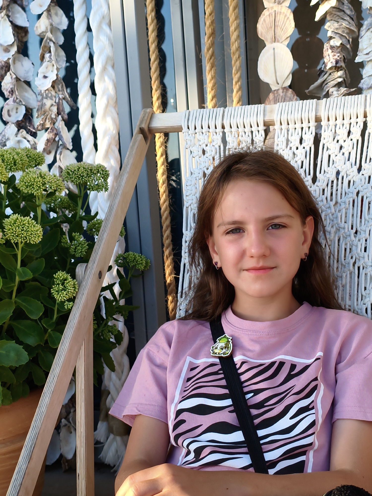 A young girl with long, brown hair and blue eyes is seated on a swing. She is wearing a pink t-shirt with a black and white zebra print design. A black strap crosses her chest, and a small, cartoonish pin is attached to her shirt. She is looking at the camera with a slight smile. The swing is made of white macrame material and has wooden armrests. Behind the girl, there are ropes, shells, and plants. The scene appears to be outdoors, possibly on a patio or deck, with a blurred background suggesting a city environment.