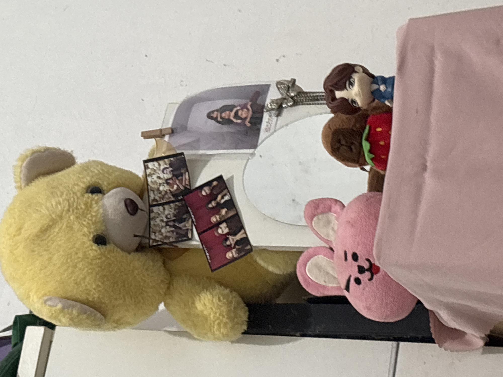 In the city of North Jakarta, Indonesia, a bedside shelf displays several items. A large yellow teddy bear is positioned on the left, holding two small framed photographs. To its right, another framed photograph is pinned to the wall, showing a group of figures stacked vertically. Below these, a pink plush rabbit with a drawn-on face peeks out from behind a pink cloth. Next to it, a brown plush strawberry and a small figurine of a person in a blue shirt are visible. The scene is lit by artificial light, casting shadows on the white wall behind the objects.