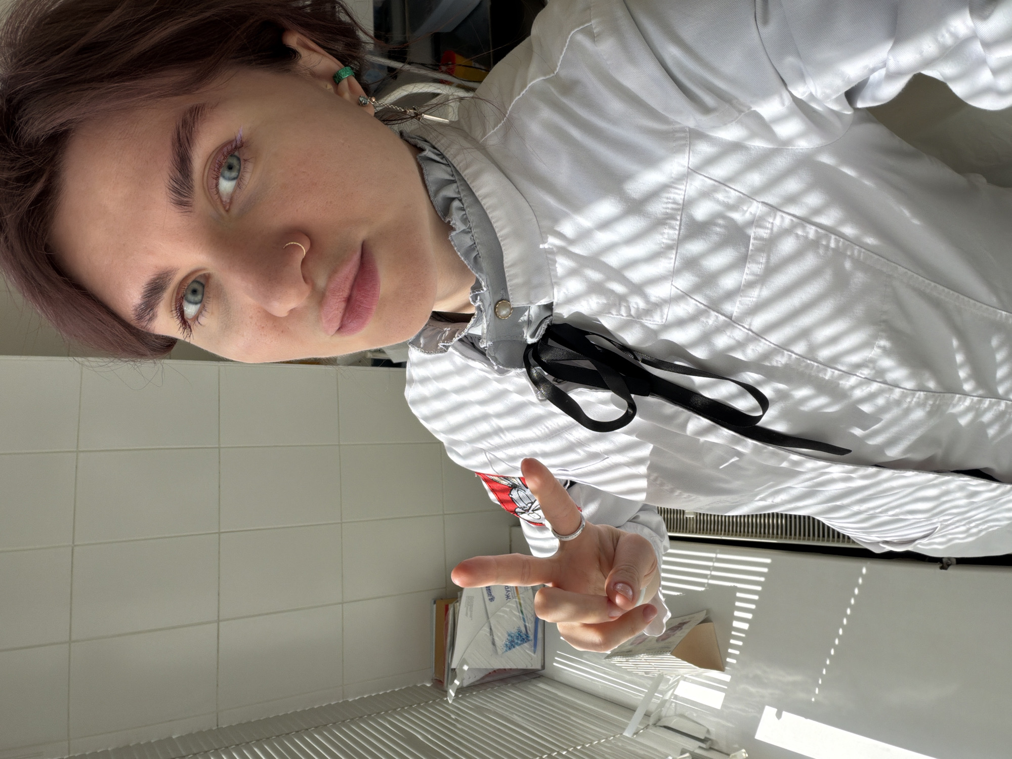 A person is visible in an interior setting, angled from the viewer's perspective. The person has fair skin, light blue eyes, and dark brown hair. A small gold-colored hoop is visible in their left nostril. They are wearing a white shirt with a gray ruffled collar and a black ribbon tied at the neck. The shirt appears to have a pocket. A hand is in the foreground, displaying a ring on one finger. The background shows a white tiled wall on the left. Beneath the person, there's a radiator and some packages on the windowsill.
There is no indication in the image that it was taken in Khimki, Russia.
