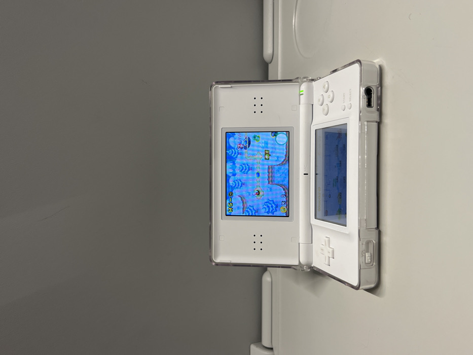 A white Nintendo DS Lite handheld gaming console, encased in a clear protective shell, is displayed in a vertical orientation. The console is open, with its two screens facing the viewer. The top screen is actively displaying a colorful video game, featuring a blue, aquatic or sky-themed level with various enemies and platforming elements. Visible text on the game screen includes "W5-SM" (likely indicating World 5-Mini or a similar level designation), "03" coins, and "0x" lives.
The bottom screen of the console appears dark or off, reflecting a faint blue hue. The console's buttons are visible, including the D-pad, face buttons (A, B, X, Y), and "SELECT" and "START" buttons. A green indicator light is illuminated near the top right of the console, suggesting it is powered on.
The background consists of two distinct vertical surfaces. To the left of the console is a dark grey wall or panel, which shows some subtle scratches or marks. To the right, and partially behind the console, is a lighter, off-white wall or vertical structure, possibly a door frame or piece of furniture. The console itself appears to be propped against or hanging from this lighter surface. The overall lighting suggests an indoor setting.