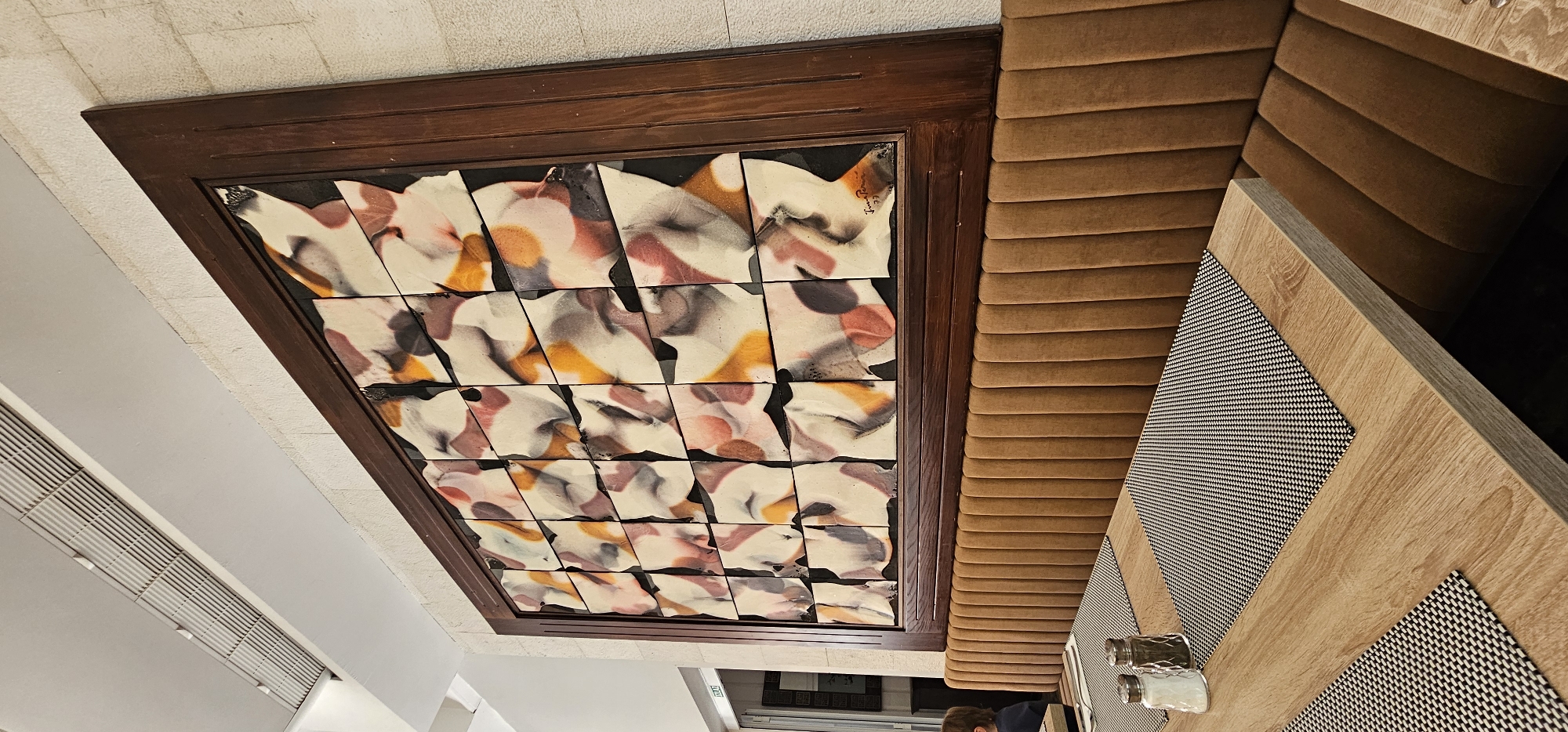 This image shows a section of a restaurant's interior in Grad Supetar, Croatia. The foreground features a light-colored wooden table with two black and white checkered placemats. On one placemat are salt and pepper shakers. A person with dark hair is partially visible sitting at the table on the left. Behind the table is a long, tufted brown velvet bench seat. Above the bench seat, mounted on a light-colored textured wall, is a large, framed piece of art. The artwork is composed of numerous square tiles with a marbled, abstract design in shades of white, pink, brown, orange, and black. The frame is dark wood. Above the artwork is a white ceiling with a large air vent grille. The overall impression is one of a modern, clean dining space.