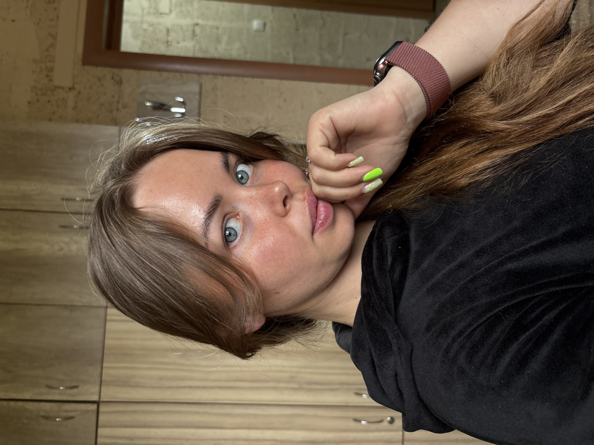 A woman is lying down, facing the camera, with her hand near her face. She has light brown hair and blue eyes, and is wearing a black velour top and a wristwatch with a pink mesh band. Her nails are painted neon green. The background appears to be a wooden cabinet with drawers and a framed mirror on the wall. The location is Русский Вожой, Russia.
