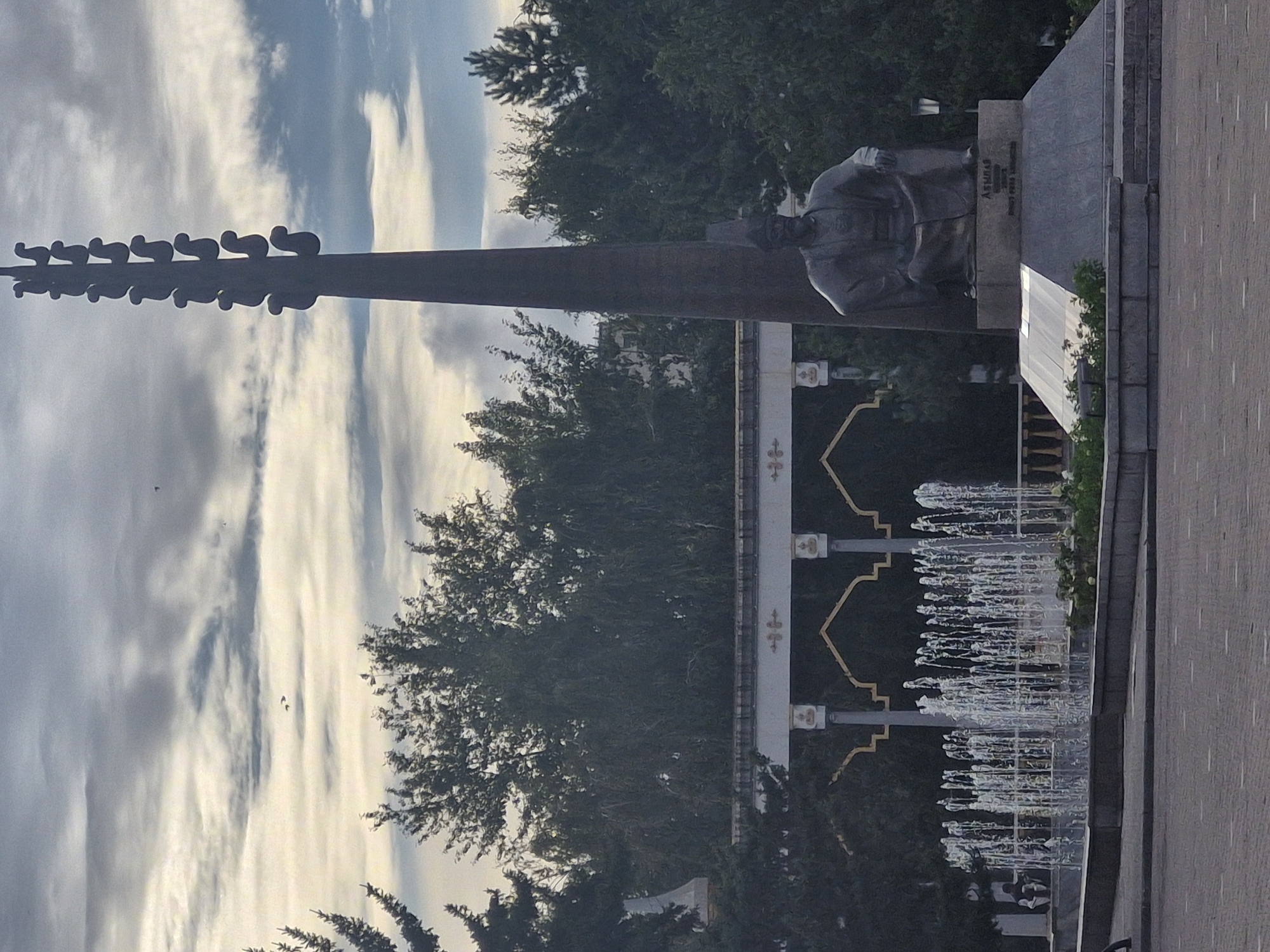 The image depicts a monument located in the city of Kokshetau, Kazakhstan, likely in a public square or park. In the foreground, a large stone monument features a seated statue of a man wearing traditional attire and a tall, tapering column adorned with a series of sculpted figures at the top. To the left of the monument, a fountain is active, with numerous jets of water creating a shimmering display. Behind the fountain and monument, an arched structure with decorative elements is visible, and dense green trees form a backdrop against a cloudy sky. The ground in the foreground is paved with interlocking bricks. The scene appears to be a public space, possibly a landmark or memorial within the city.