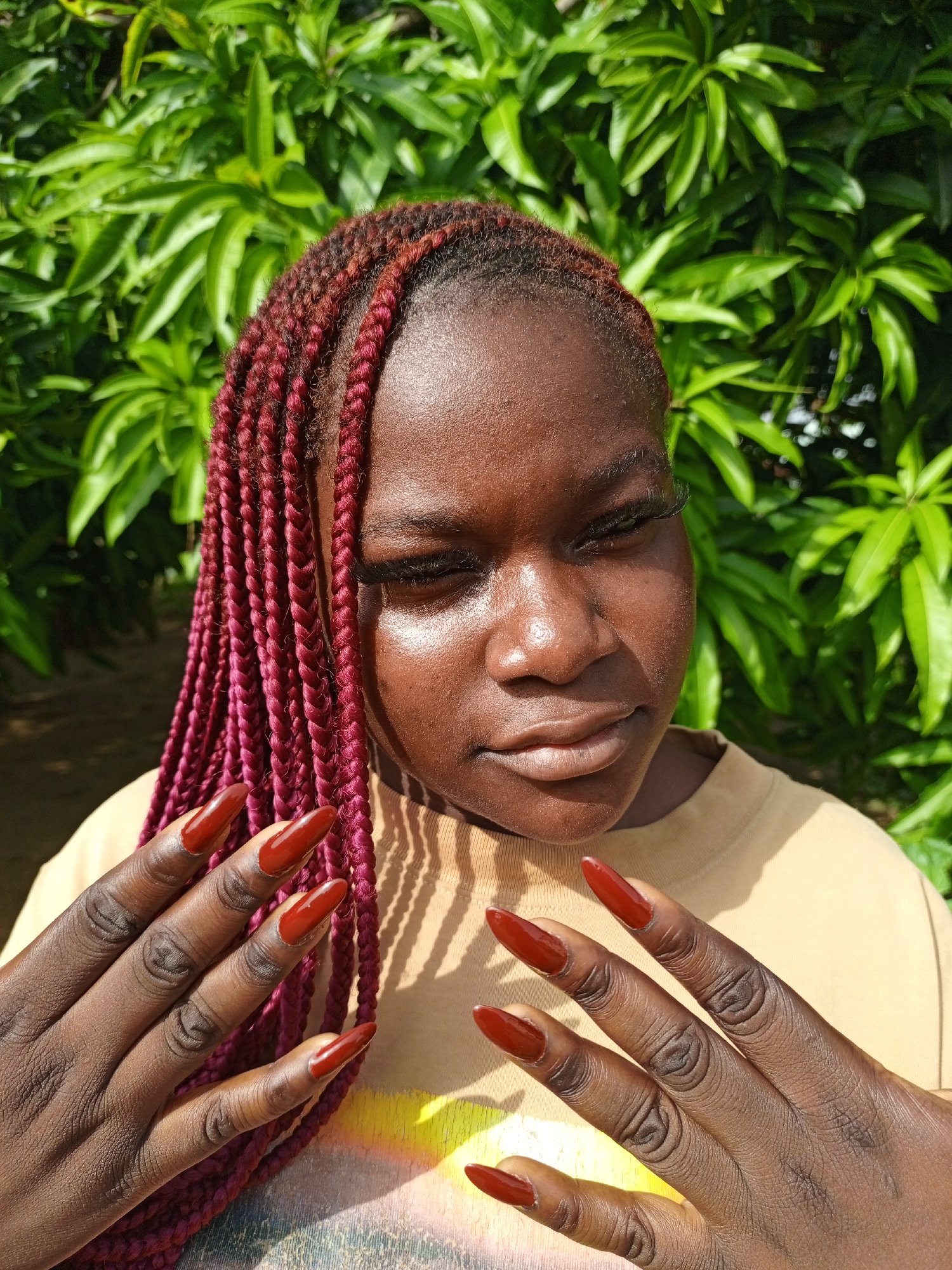 A close-up outdoor portrait features a young person, likely female, with dark skin, posing against a backdrop of vibrant green foliage under bright sunlight. Her long, reddish-maroon box braids are styled with a side part, framing her face. Her eyes are partially closed, and her expression is neutral, possibly due to the intense midday sun which casts strong highlights on her forehead, nose, and cheeks. She wears a light tan t-shirt with a faint, illegible graphic design on the chest. Both of her hands are raised, prominently displaying long, pointed, dark red artificial nails. The lush green leaves in the background suggest a natural, outdoor environment, consistent with the provided context of Keffi, Nigeria. The image focuses on the subject's distinctive hairstyle, intricate nail art, and the effect of natural light on her skin.