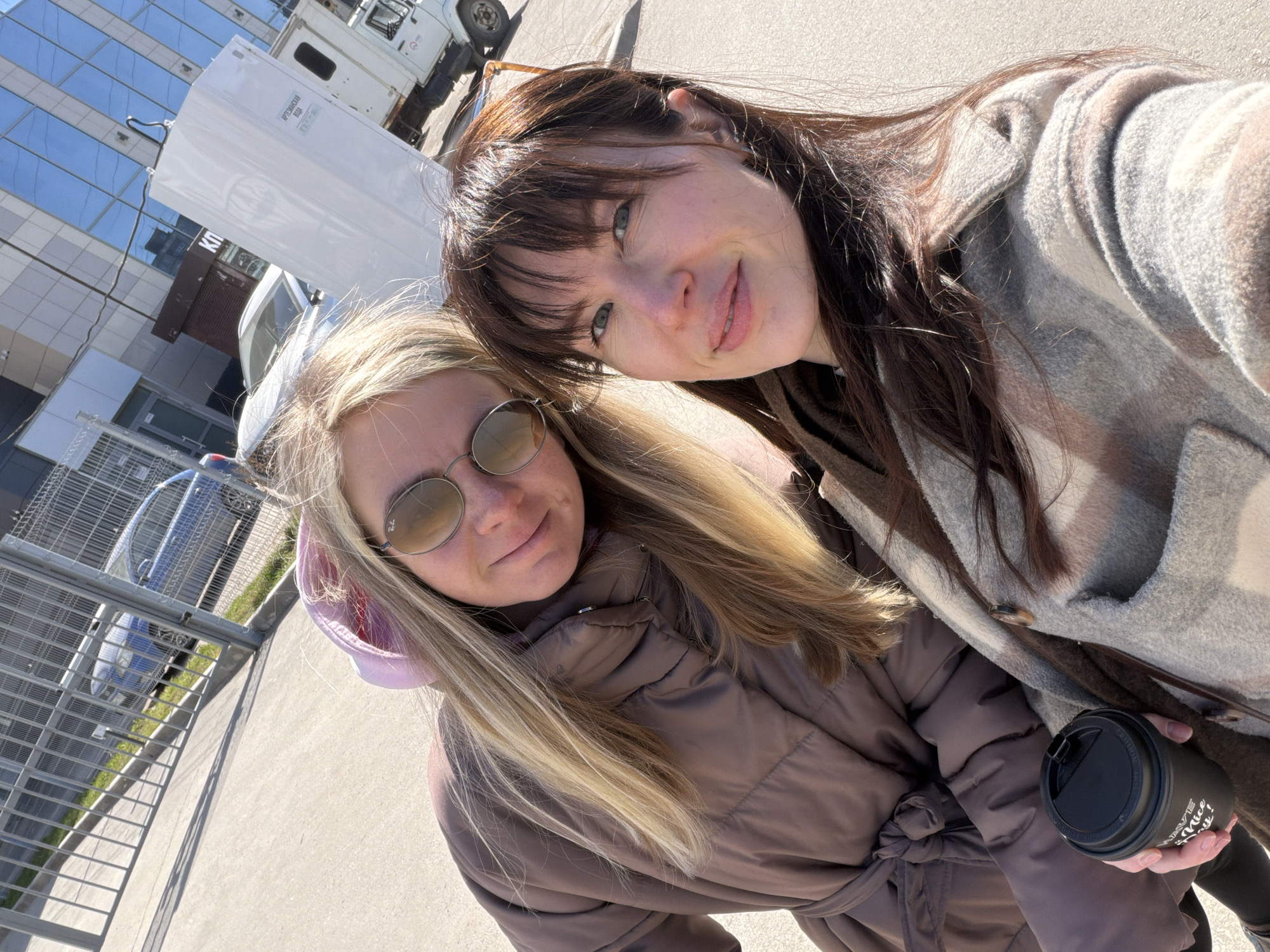 Two women are taking a selfie outside on a sunny day in Perm, Russia. The woman on the left has blonde hair, is wearing sunglasses, and a brown puffer jacket with a pink hoodie underneath. The woman on the right has dark hair with bangs and is wearing a gray and brown coat, holding a black coffee cup. Behind them, there is a large modern building with mirrored windows and a white truck with a sign in Russian on the side, indicating "Artesian Water". A blue shopping cart and some metal fencing can be seen to the left, indicating a commercial or industrial area. The ground is concrete, and the angle indicates the photo was taken from a slightly high perspective.