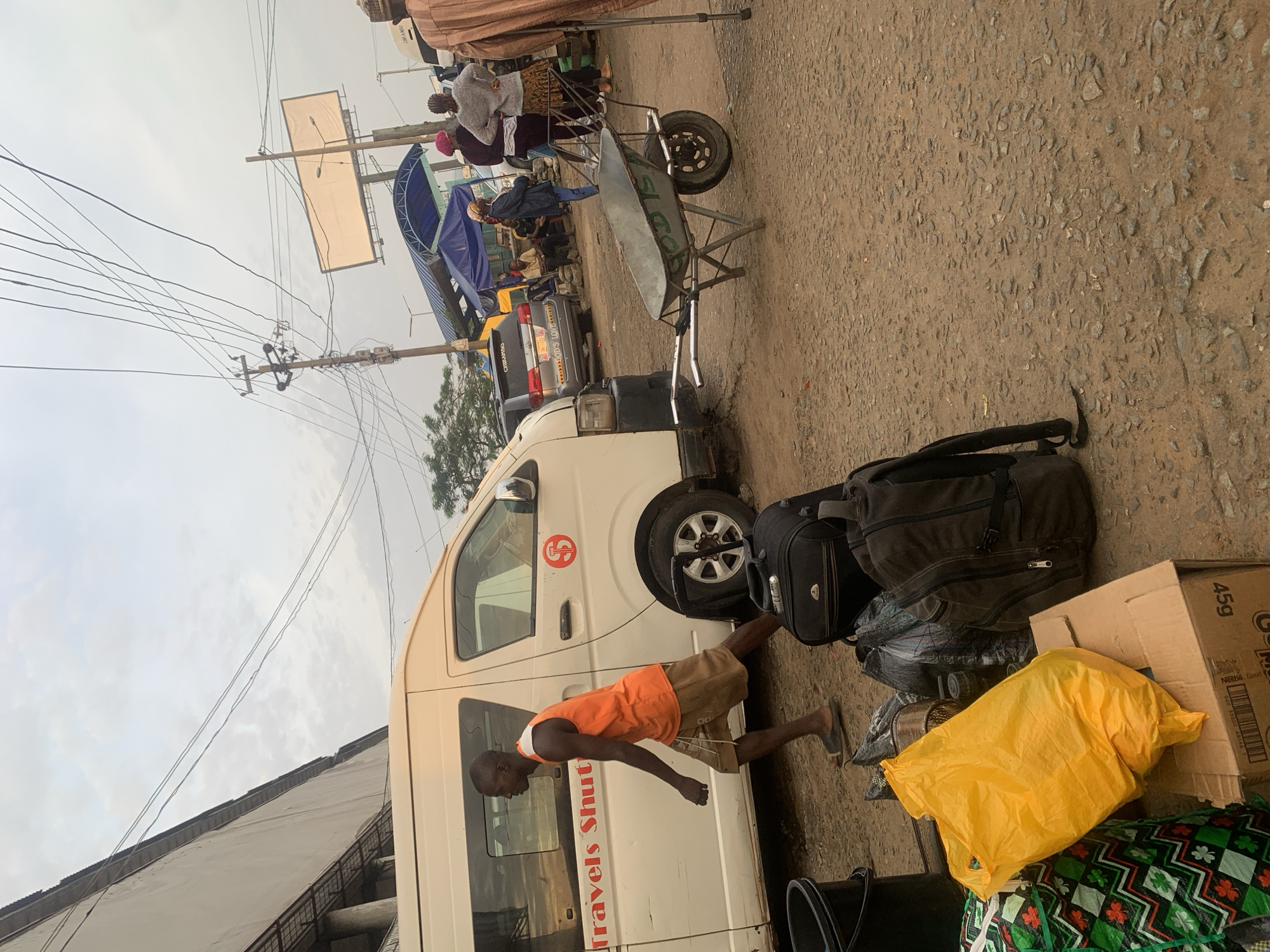 In Elelenwo, Nigeria, a street scene unfolds under a cloudy sky, with a white van labeled "Travels Shut" parked on the left. A Black man in an orange tank top and brown shorts walks from left to right beside the van. In the foreground, several pieces of luggage are visible, including a yellow plastic bag, a cardboard box, and two backpacks. A wheelbarrow, marked with "GOD IS," is positioned in the middle of the dirt and gravel road. Further in the background, several people are gathered near stalls and another vehicle. Overhead, a network of electrical wires crisscrosses the sky, and a large, blank billboard stands on a pole. The environment appears to be a bustling outdoor market or transport hub.