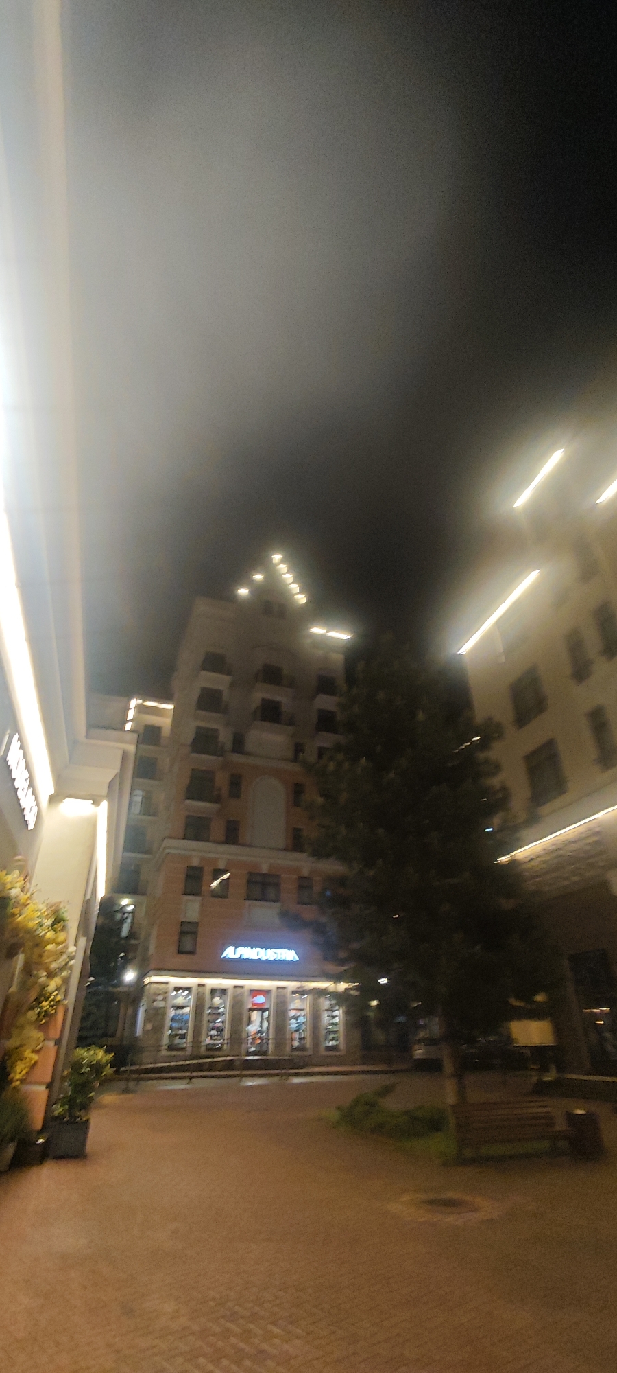 The image depicts a nighttime scene in Estosadok, Russia. A multi-story building with the sign "ALPINDUSTRIA" is visible at ground level. The building is peach-colored with multiple balconies and decorative lights on its roofline. A large tree partially obscures the view of the building. To the left and right, portions of other buildings are visible. The ground is paved with brick-like tiles. There are no people visible in the scene. The lighting suggests a commercial or public area.