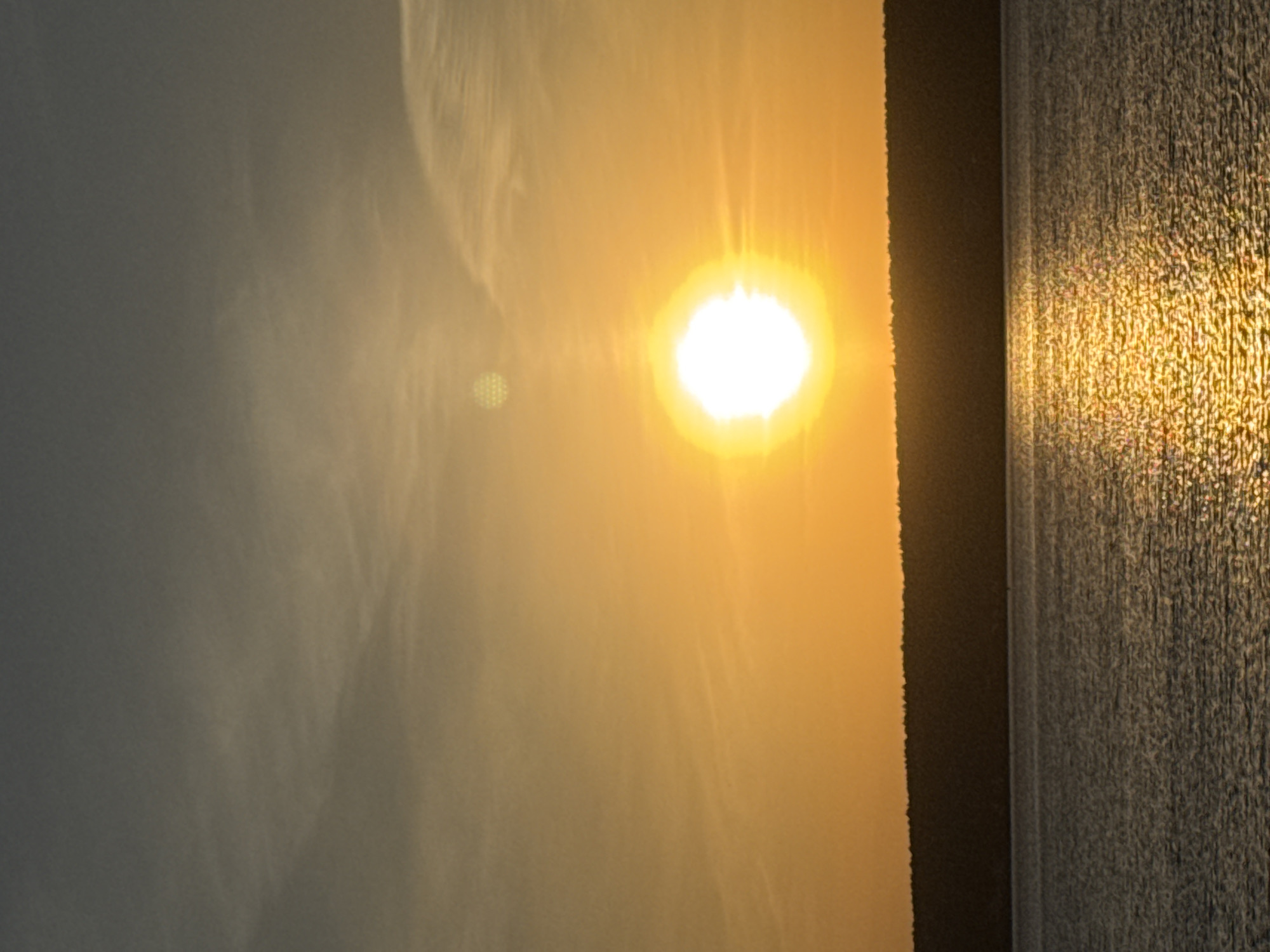 The image shows a bright sun shining through what appears to be a window. The light illuminates a textured surface or material on the right side of the frame, while a grayish area fills much of the left. The sun itself is positioned slightly off-center, creating a distinct glare. The setting seems to be indoors. There is no visible evidence to pinpoint this scene in Naberezhnye Chelny, Russia.