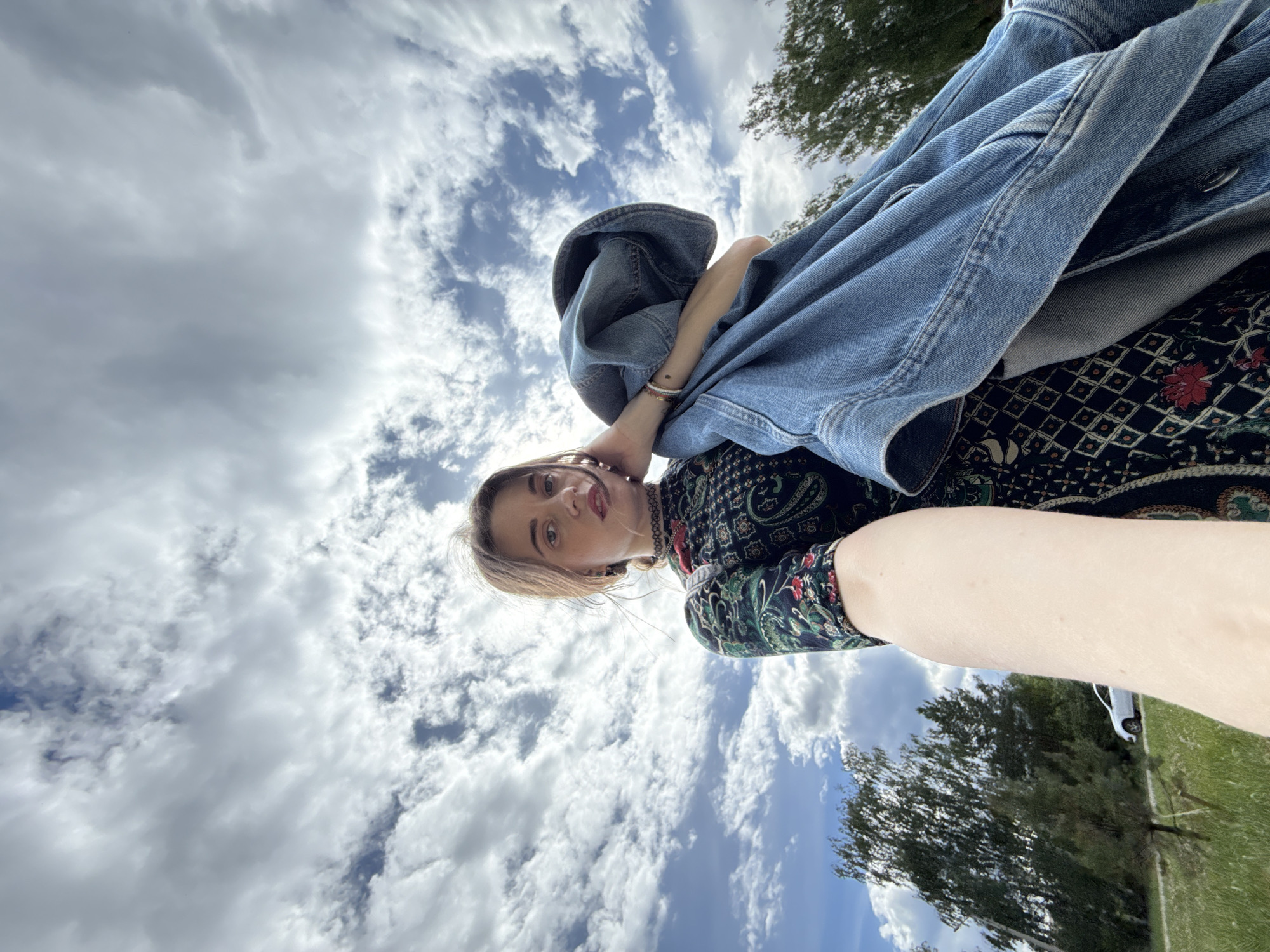 A woman is photographed from a low angle, holding a denim jacket in front of her chest. She is wearing a dark patterned dress and a black choker necklace. Her right hand is touching her face. The background shows a cloudy sky with patches of blue, trees, and a white car on a road.