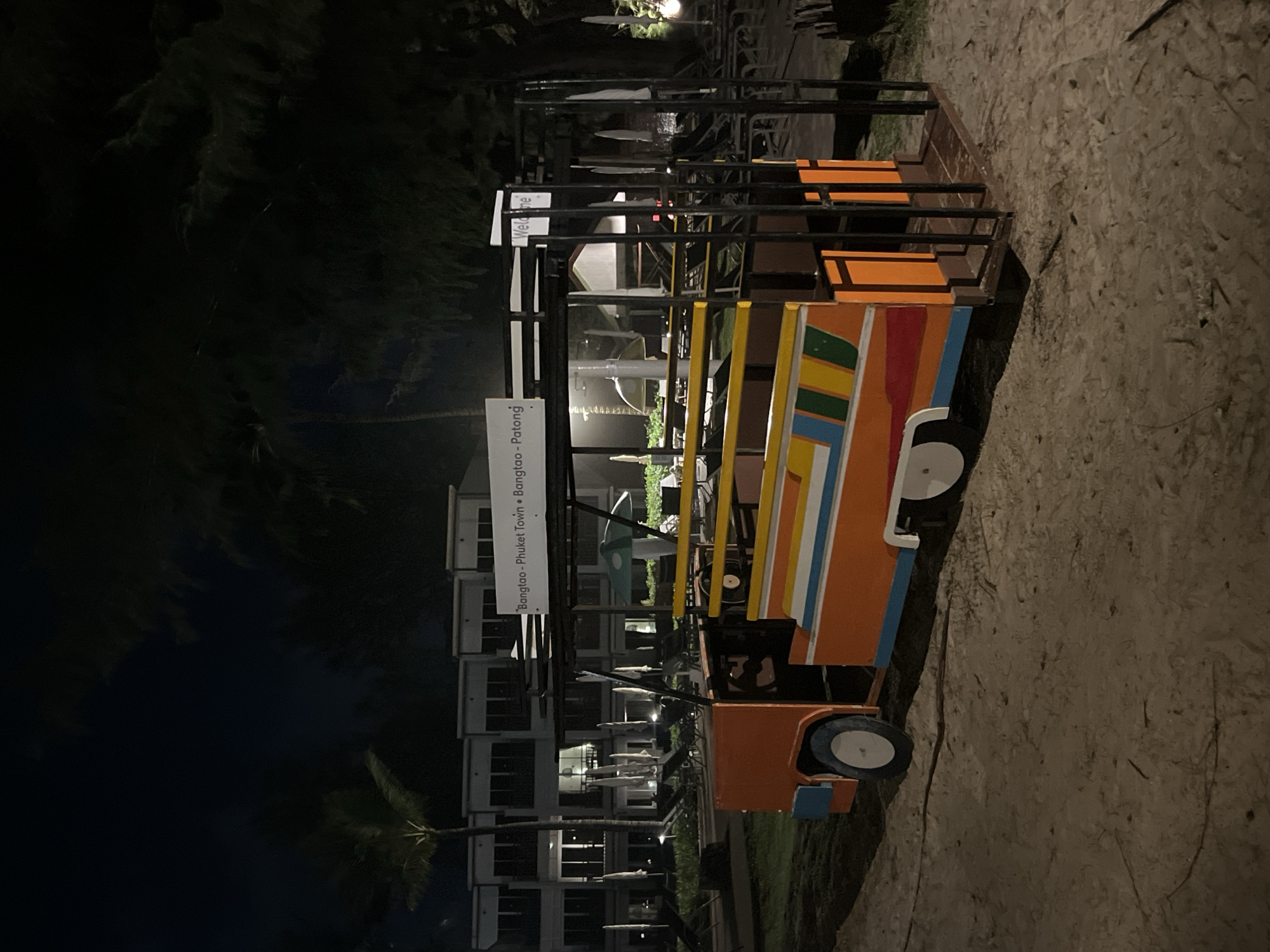 This is a night-time photograph taken outdoors, likely in a tropical location given the presence of palm trees. The scene features a brightly colored, multi-passenger vehicle that appears to be a tuk-tuk, a common mode of transport in Southeast Asia. The tuk-tuk has an orange base with colorful stripes of yellow, blue, green, and red along its side. It has a metal frame structure above for passengers, and sits on a sandy or gravelly surface.
In the background, there is a modern building complex with multiple floors and windows, suggesting a resort, hotel, or residential area. The lighting is dim, indicating it is nighttime, and artificial lights from the building and possibly the tuk-tuk itself are visible. The atmosphere is dark, with shadows cast by the surrounding environment, including dense foliage that looks like palm trees.
There are two visible signs attached to the tuk-tuk's frame. One sign, in white with black text, appears to read "Welcome". Below it, another sign with white text on a dark background reads "Bangtao - Phuket Town • Bangtao". This text provides specific location cues, indicating that the scene is likely in Bang Tao, Phuket, Thailand, and that the vehicle serves routes to Phuket Town.
No people are clearly visible in the frame. The overall mood is quiet and still, characteristic of a peaceful night in a tourist destination. The weather appears to be clear, as there are no indications of rain or strong winds.