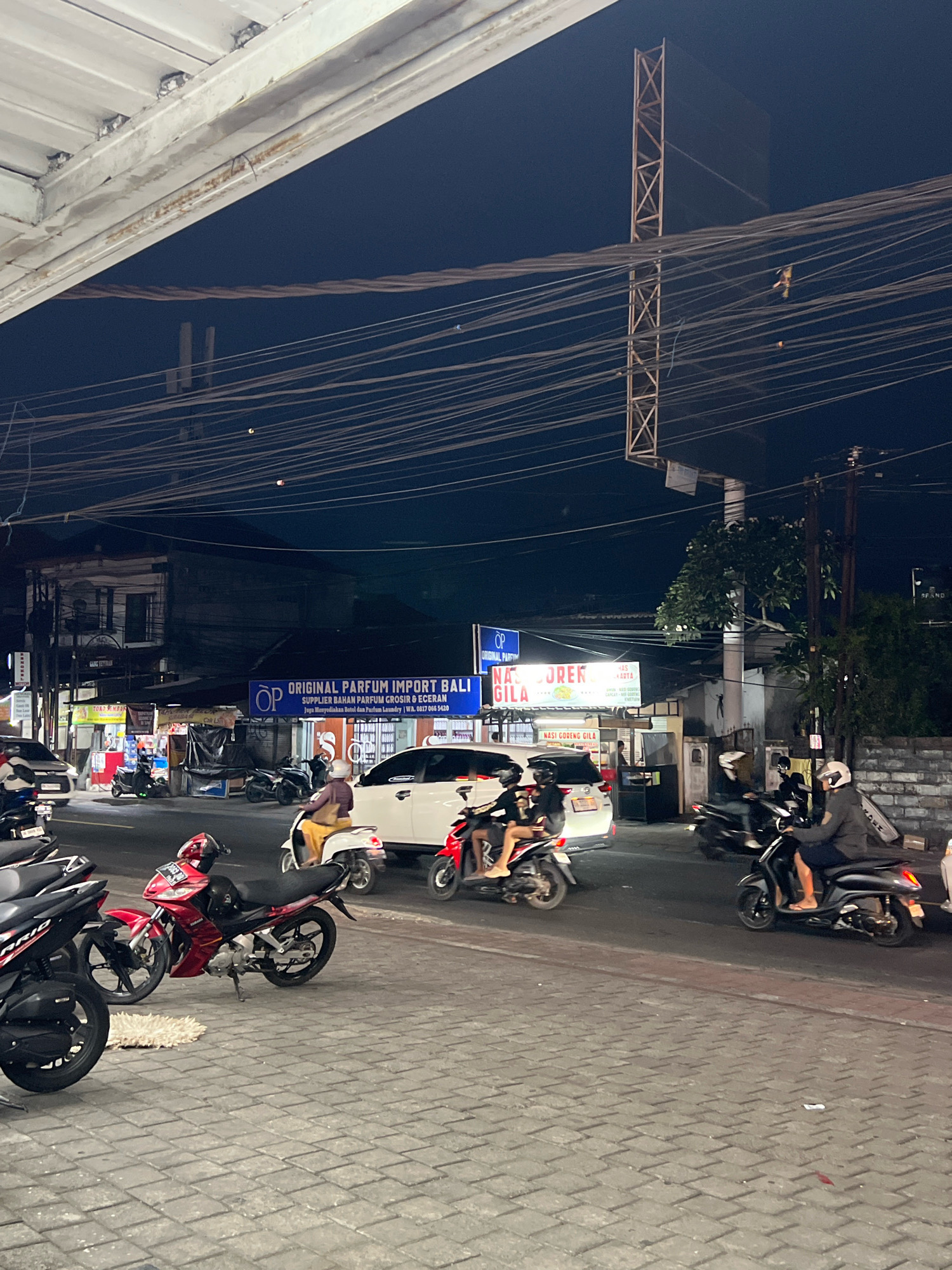 The image depicts a street scene at night in Denpasar, Indonesia, likely near a commercial area. The foreground shows a brick-patterned sidewalk with several motorcycles parked and in motion. A red and black motorcycle is prominently displayed, parked at an angle. Further down the street, a white car is visible, with people on motorcycles around it. Signs for "ORIGINAL PARFUM IMPORT BALI" and "NASI GORENG GILA" are illuminated, indicating businesses in the vicinity. Numerous overhead wires crisscross the dark sky, typical of urban infrastructure. The overall atmosphere is that of a busy street at night, with traffic and businesses operating.