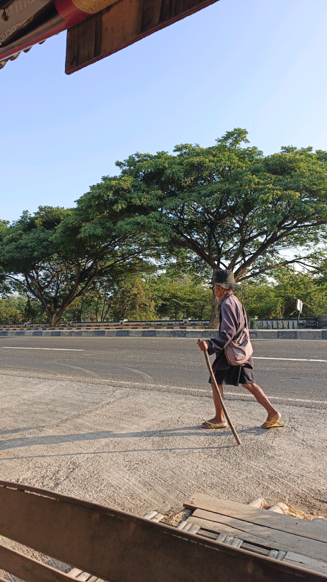 An elderly man with a cane is walking on the sidewalk next to a road. He is wearing a hat, a long-sleeved shirt, and shorts. There are large trees in the background. The scene appears to be in a rural or suburban area of Krimun, Indonesia. The lighting suggests it is either early morning or late afternoon. The man is casting a long shadow on the ground.
