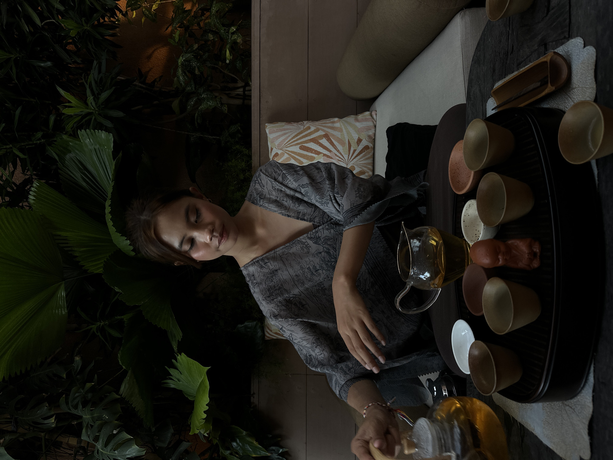 A woman is seated amongst lush greenery, preparing tea. She is wearing a gray, patterned top and has her eyes closed, appearing relaxed. In front of her is a tea set with various cups and a glass teapot filled with tea. She is pouring tea from one teapot into another. The setting appears to be an indoor space with outdoor plants integrated into the design. The location is Pecatu, Indonesia.