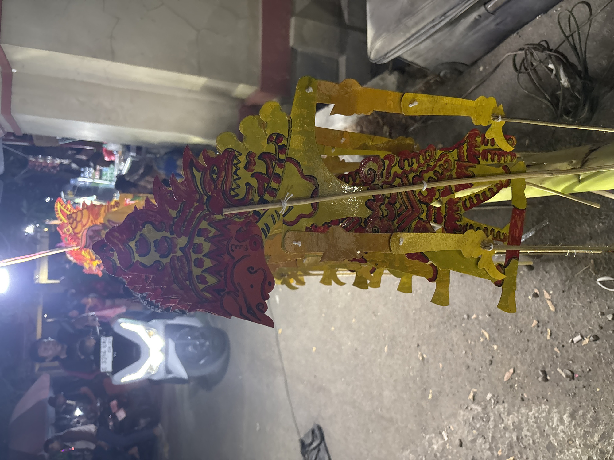 In Panongan, Indonesia, a line of traditional Wayang Golek puppets, characterized by their red and yellow designs, stand upright on wooden sticks. In the background, a person rides a white scooter, its headlights illuminated, on a street. Several other people and a red umbrella are also visible in the background, suggesting a bustling street scene or a local event. The ground is paved with asphalt, and debris, including leaves and small stones, is scattered across it. A metal object, possibly a container, is present in the lower right corner of the image.