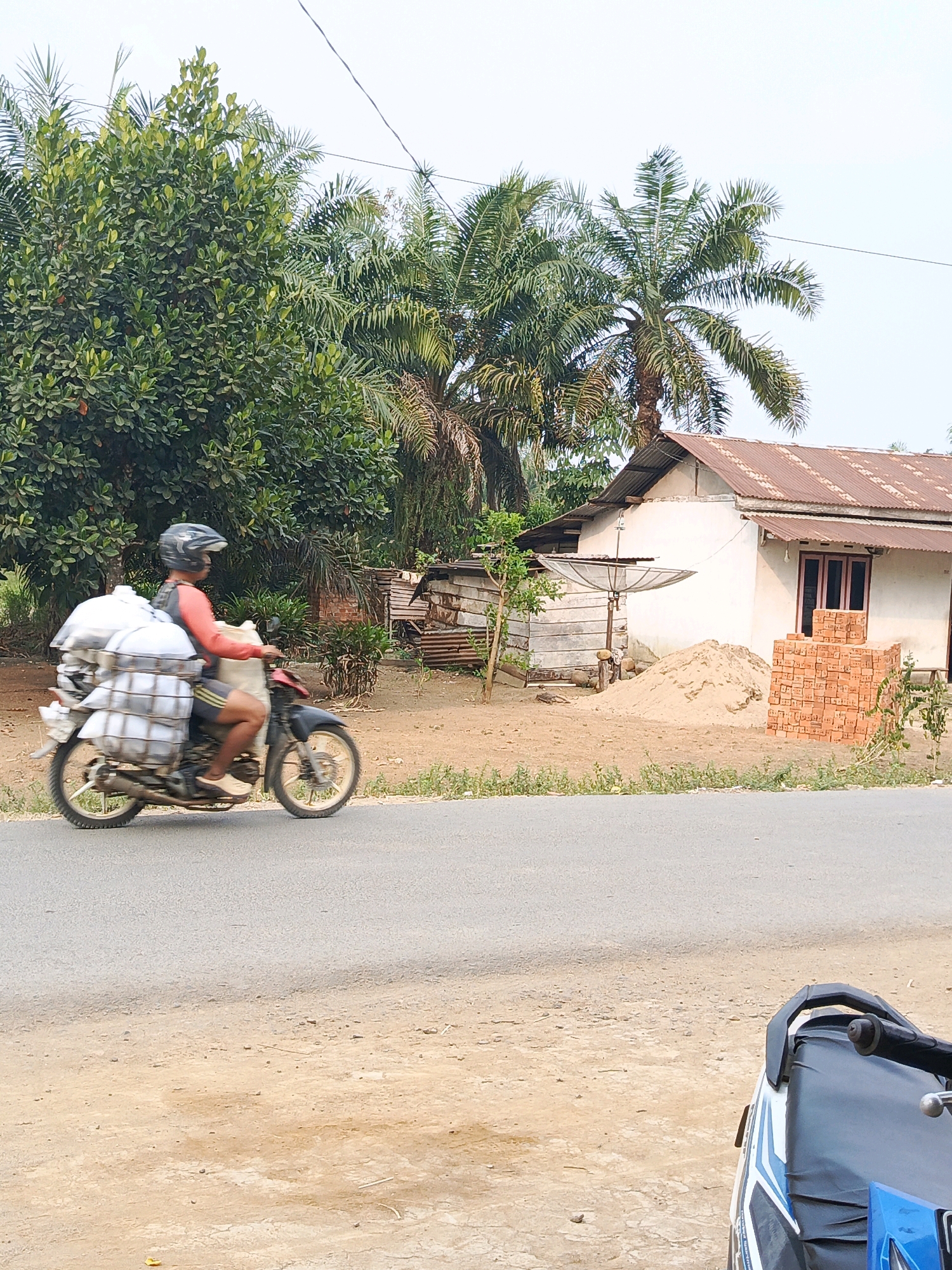 A person wearing a red shirt, dark shorts, a helmet, and light-colored shoes is riding a red and black motorcycle on a paved road. The motorcycle is carrying a large load of white sacks secured to the rear. The scene appears to be in an outdoor, rural or semi-rural area, likely Megang Sakti I, Indonesia, given the context. Behind the motorcycle, there is a house with a rusted tin roof, a pile of sand, and a stack of red bricks. Lush green trees and palm trees are visible in the background, with a clear sky overhead. The road surface is asphalt, and the ground beside the road is a mixture of dirt and sparse vegetation.
