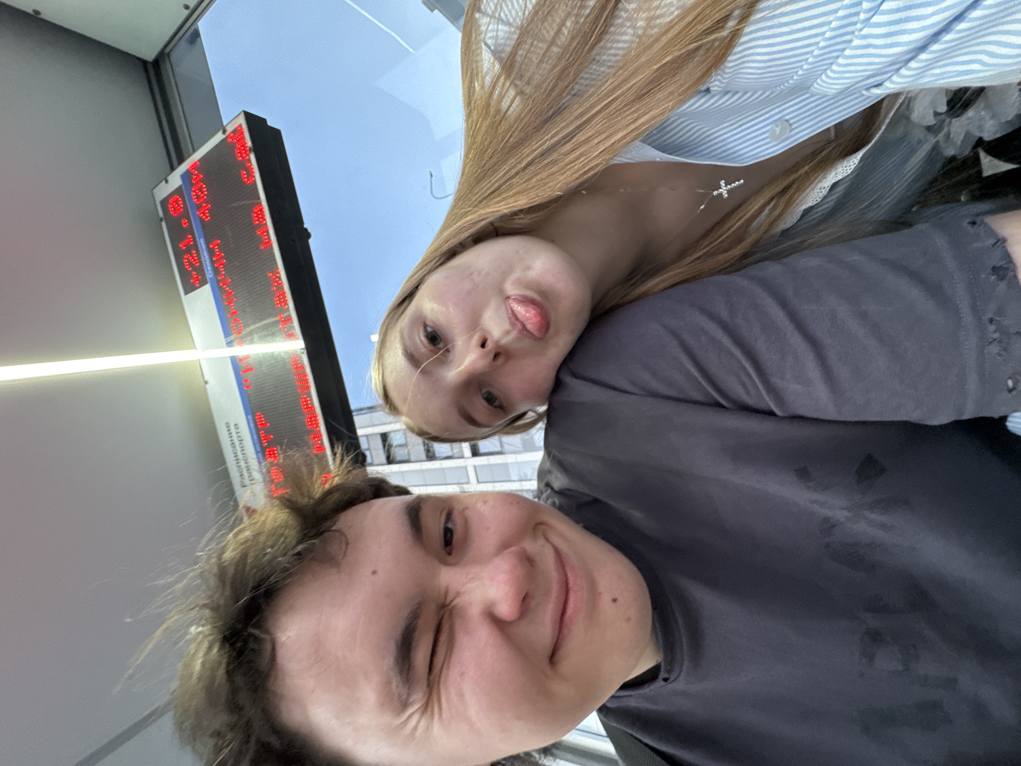 Two individuals, a male and a female, are positioned closely together, apparently taking a selfie. The male, in the foreground, is winking. The female, slightly behind, is sticking her tongue out. Both are wearing casual clothing. In the background, partially obscured, is an LED display showing what seems to be transit information in Russian. The sign contains the words “Расписание” and “транспорта”, suggesting it may be in a bus or train station. Based on the font and other contextual clues, it is highly probable that this is located in the city of Perm, Russia.