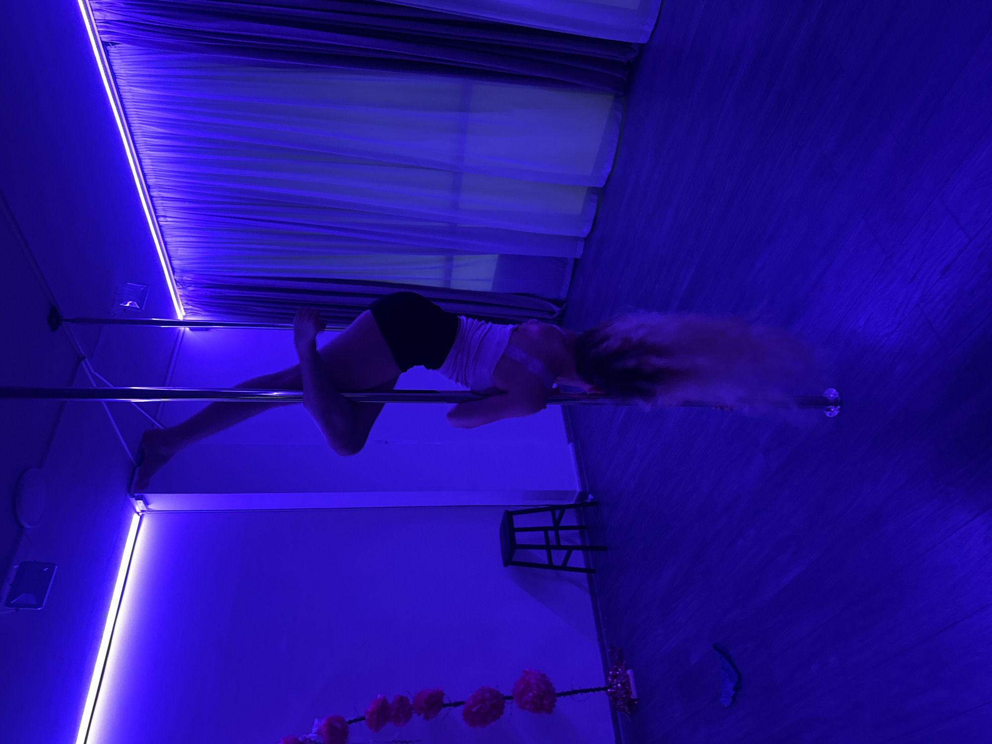 This media file captures an indoor scene dominated by deep blue lighting, creating a dramatic and atmospheric setting. The central subject is a person, appearing to be a woman with long, flowing light-colored hair, actively engaged in pole dancing. She is shown in an athletic, inverted pose on a metallic chrome pole, with her body curved and one arm and leg gripping the pole. She is dressed in a light-colored top and dark shorts.
The setting is likely a dance or fitness studio. The walls, though possibly light-colored, are bathed in the blue ambient light. Contrasting with this are bright white LED strip lights, one running horizontally along the top of the wall near the ceiling and another vertically along a wall segment to the left. In the background, windows are covered by blinds or curtains, also illuminated by the blue light. The floor is made of dark wood.
Additional objects visible include a black, open-frame stool positioned on the right side, and a garland of pink or red artificial flowers strung horizontally across the lower part of the scene. The intense artificial blue and white lighting suggests the activity is taking place at night or in a controlled studio environment, rather than under natural daylight. No visible text is present in the image.