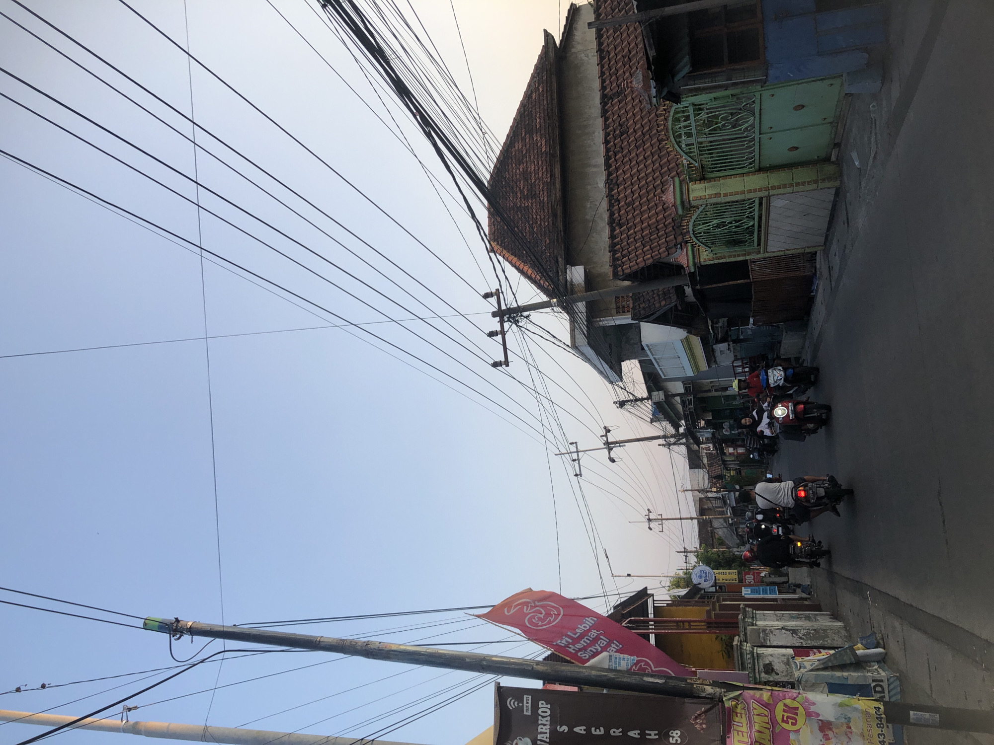 The image depicts a street scene in Tenggulunan, Indonesia, characterized by a clear sky and numerous overhead power lines. The street is paved and appears to be a typical urban or suburban thoroughfare. Several motorcycles are in motion, with riders navigating the road. Buildings line the street, some featuring traditional tiled roofs. A red flag with a white logo is prominently displayed, attached to a pole. Signs are visible on some buildings, indicating commercial establishments. The overall atmosphere suggests a normal day with daily activities underway.
