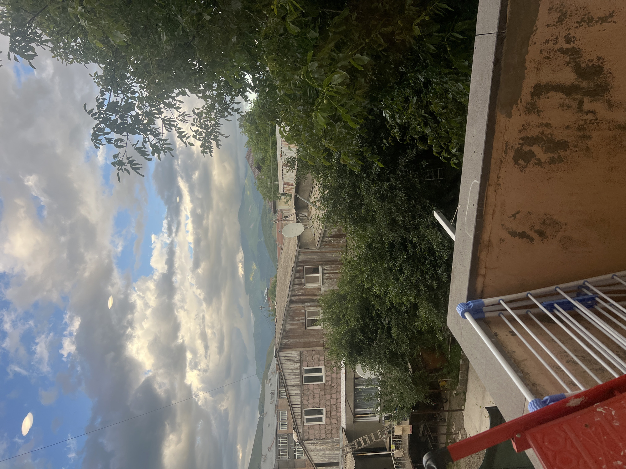 The image depicts an outdoor scene, likely in a residential area, with a building and vegetation in the foreground and midground, and mountains and sky in the background. The perspective is from a high vantage point, looking down.
In the foreground, on the right, there is a concrete balcony railing with a white drying rack attached, some of which is obscured by a red object. Lush green trees with dense foliage dominate the left and mid-right portions of the image, partially obscuring the view of the buildings and the landscape beyond.
The midground features a multi-story building with a weathered, rustic appearance. The upper portion of the building has a corrugated metal roof and is constructed with wooden planks. The lower portion appears to be made of stone or brick. Several windows are visible on the building's facade, some with white frames. A satellite dish is mounted on the roof. Next to the building, more trees are visible. In the distance, rolling hills and mountains are partially obscured by haze and trees.
The sky in the upper left and background is a mix of blue sky and dramatic, heavy clouds. The lighting suggests it is either early morning or late afternoon, with soft, diffused light. The overall mood is somewhat serene, with a hint of rustic charm from the weathered building and the natural surroundings. There is no visible text in the image. The context provided places this scene in Vanadzor, Armenia.