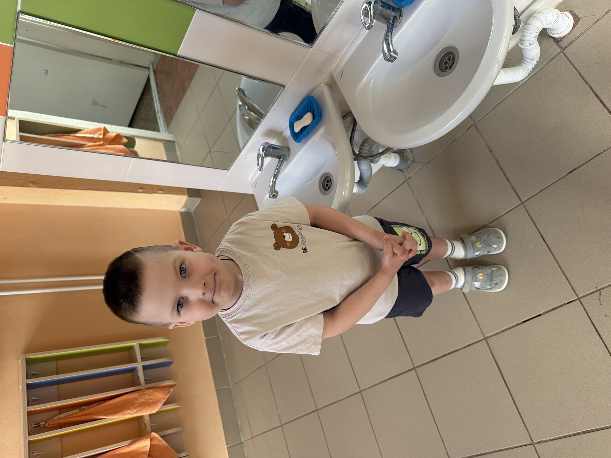 This media file captures a young boy standing in what appears to be a children's washroom or bathroom, located in Chaikovsky, Russia. The boy, who looks to be a toddler or preschooler with light hair cut short and bright blue eyes, is centrally positioned in the frame, looking towards the camera with a soft, gentle smile. He is wearing a light beige t-shirt featuring a brown cartoon bear graphic and the text "BE AWESOME" printed alongside it. He also wears dark shorts and grey closed-toe sandals over white socks, with his hands clasped together in front of him.
The setting is designed for children, featuring two white sinks with chrome faucets, one slightly higher than the other, to the boy's right. A blue soap dish holding a bar of white soap is placed between the sinks. Mirrors are mounted above the sinks, reflecting parts of the room, including a glimpse of orange towels. A white flexible drain pipe is visible beneath one sink. The floor is covered in light beige square tiles, and the walls are a mix of light orange paneling and green, white, and orange tiles. On the left side of the image, a wooden rack with colorful slots (green, blue, and orange) holds neatly folded orange towels, suggesting a designated spot for each child's towel. The environment appears clean and functional, indicative of a daycare, preschool, or kindergarten facility. The lighting is internal, offering no specific cues about the time of day or weather.